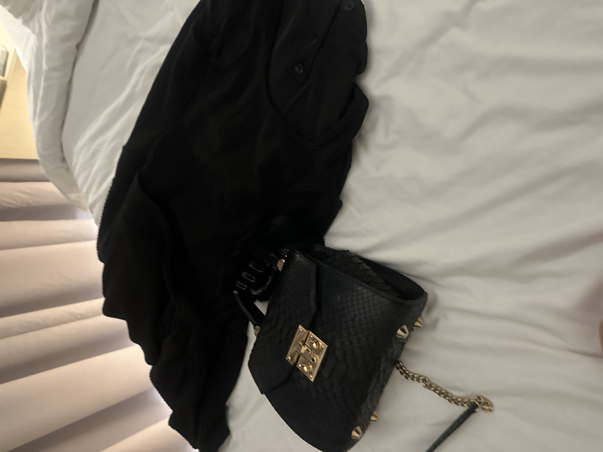 A black textured handbag with gold hardware and spikes rests on a white duvet. A black garment, possibly a sweater or dress, is draped over the duvet behind the handbag. The background consists of light-colored vertical curtains. The scene is set indoors, on a bed, in North Kuta, Indonesia.