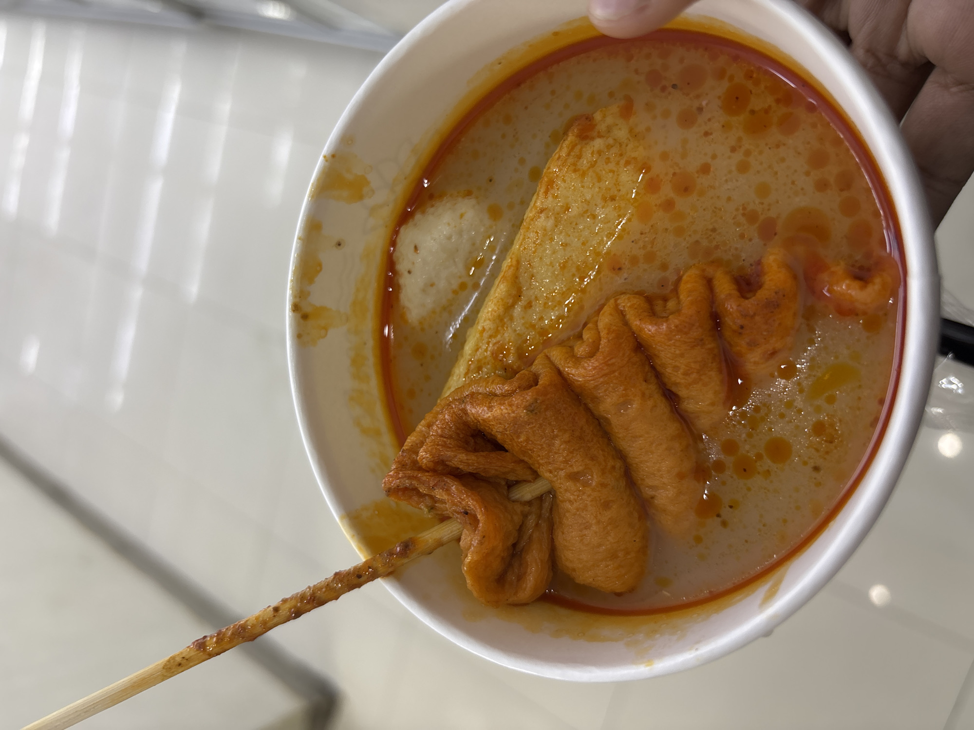 This close-up image displays a white disposable bowl filled with a savory, milky-orange broth, held by a person's hand at the top right. The broth's surface is dotted with oil droplets, and a deeper reddish-orange hue rings the inside of the bowl's rim, suggesting a spicy or seasoned flavor.
The bowl contains several pieces of orange-brown fish cake (eomuk or odeng). A large, rectangular piece is partially submerged, while another is folded onto a wooden skewer, typical of Korean street food. The skewer extends out of the bowl towards the bottom left. A smaller, lighter-colored, possibly white fish ball or similar item is also visible within the broth.
The background features a light-colored, reflective surface, likely a tiled or polished floor, showing bright, elongated reflections of overhead lighting. This setting suggests an indoor public area such as a food court, market, or convenience store, commonly found in urban environments like West Jakarta, Indonesia, where such street food is popular. No text is visible in the image.