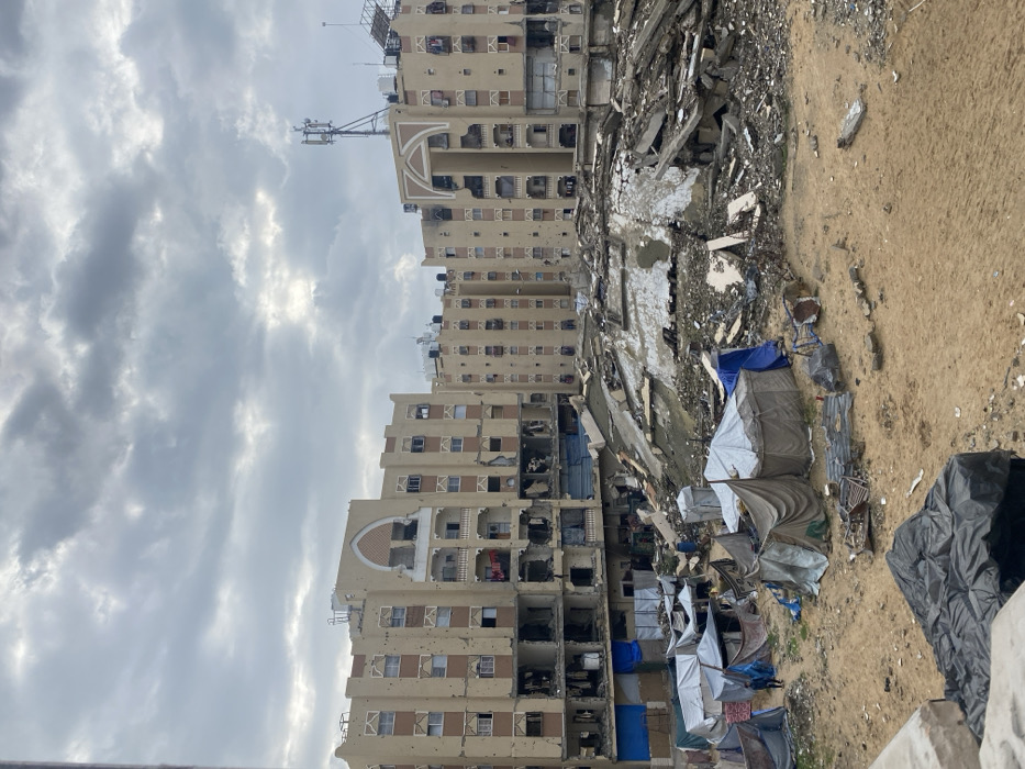 The image displays a severely damaged urban environment in Khan Yunis, Palestinian Territory, under an overcast sky. In the background, multiple multi-story buildings, primarily light brown with darker accents, show extensive structural damage. Their facades are pockmarked with numerous impact holes, windows are shattered or missing, and lower floors exhibit significant collapse, exposing interiors and creating large voids. Balconies on these buildings also show damage. A telecommunications tower is visible on the roof of one building in the distance.
In the midground, a large, irregularly shaped mound of concrete, rebar, and other construction debris occupies the space between the damaged buildings and the foreground.
The foreground consists of sandy, uneven ground where approximately 10-15 makeshift tents are erected. These temporary shelters are constructed from various materials, including white, blue, grey, and brown tarpaulins and fabric sheets, varying in size and configuration. One individual, dressed in dark clothing, is visible walking among these tents. Scattered debris, including pieces of metal, plastic, and other discarded items, lies across the sandy ground.