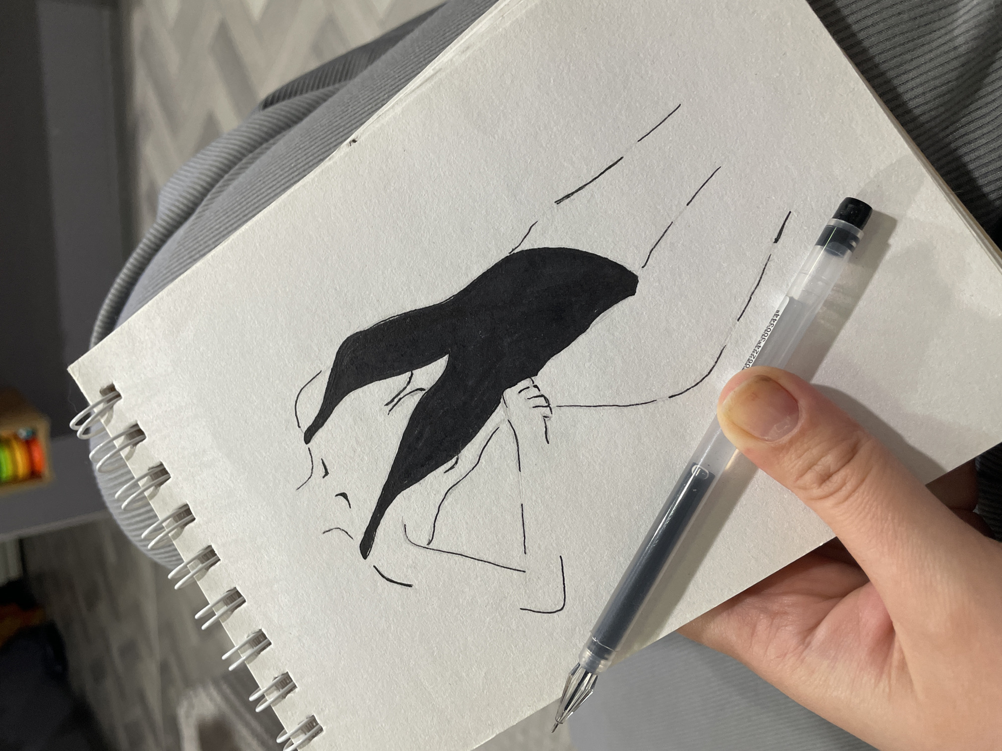 A hand holds a notebook with a drawing. The drawing depicts a figure in black ink on white paper. A black ink pen rests on the notebook. The notebook is spiral-bound. The location is impossible to determine from the image alone.