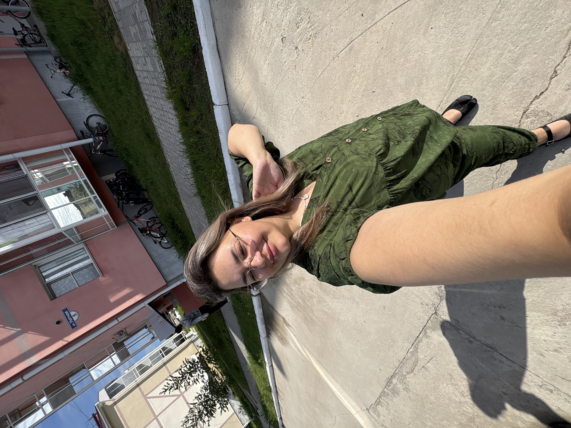 A woman stands on a concrete pavement, taking a selfie in front of a pink apartment building. She is wearing a green patterned dress and black shoes. The building has a sign that reads "ул. Уютная 8," indicating the location as Yutnaya Street, Building 8, in Birobidzhan, Russia. There are bicycles parked on the grassy area next to the building. The sun is casting shadows on the pavement.