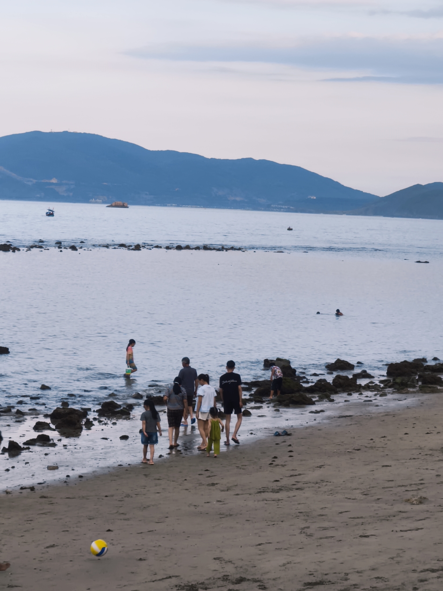 A group of people, including adults and children, are walking along a rocky beach in Nha Trang, Vietnam. The water is calm with gentle waves lapping at the shore. In the distance, mountains are visible, shrouded in a soft, hazy light. A few small boats are scattered across the sea. A yellow and white ball lies on the sand in the foreground, suggesting a casual recreational activity. The sky is a pale, overcast blue.
