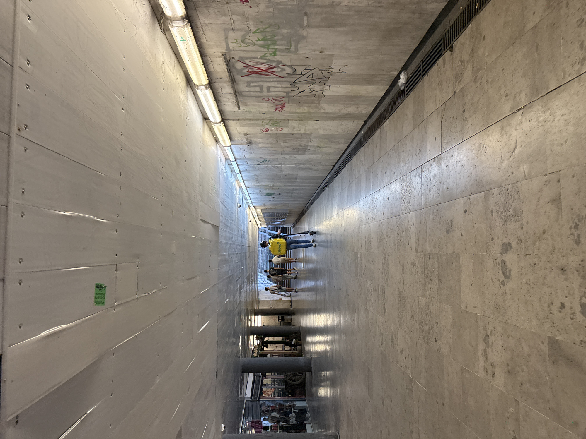 A person wearing a yellow delivery bag walks on an electric scooter through an underpass in Unknown, Unknown. The underpass has a concrete ceiling, concrete walls with graffiti, and a tiled floor. There are shops on the left side of the underpass. Several other people are walking in the distance. The lighting in the underpass is provided by fluorescent lights along the ceiling. There is a drainage grate running along the wall on the right side of the underpass.