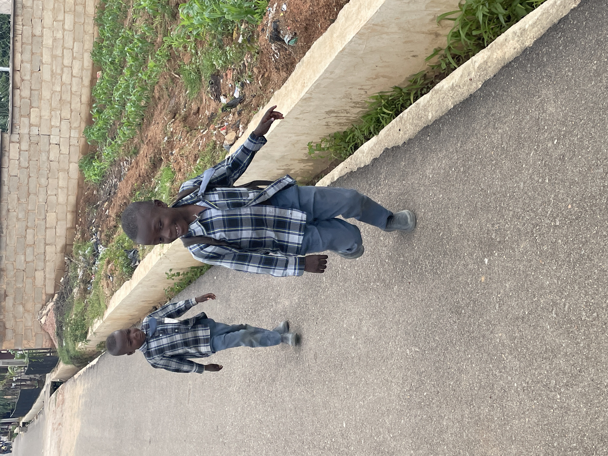 Two young boys are captured in a photograph taken outdoors in Gura Top, Nigeria. The image is oriented such that the subjects appear to be running towards the viewer on a paved asphalt surface. The boy in the foreground, dressed in a blue and black checkered shirt and grey trousers, has his mouth open in a wide smile and is looking directly at the camera with a playful expression. His right arm is extended upwards, as if in motion or reaching for something. His left arm is bent at the elbow, and he wears grey sneakers. Behind him, a second boy, also wearing a similar checkered shirt and grey trousers, is visible with his head turned towards the camera, a grin on his face. He is also wearing grey sneakers and appears to be running alongside the first boy.
The background features a steep incline covered with sparse green vegetation and some scattered debris, including plastic waste. A low, light-colored concrete barrier runs along the edge of the paved path, separating it from the vegetated area. To the left of the incline, a brick wall is partially visible. The lighting suggests it is daytime, likely with overcast conditions or dappled sunlight, as there are no harsh shadows. The overall scene depicts a moment of energetic play and joy shared by the two children in a somewhat unkempt, outdoor environment.