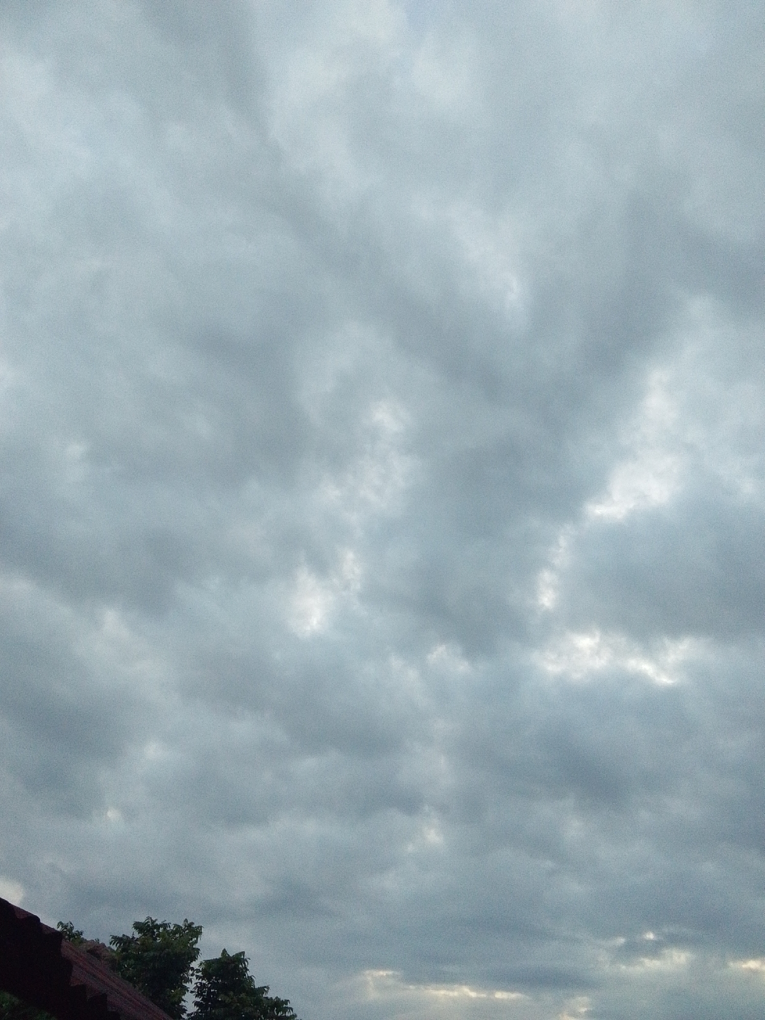 The image depicts a cloudy sky above Serti, Nigeria. In the foreground, a portion of a dark, tiled roof is visible on the left side of the frame, with lush green trees emerging from behind it. The majority of the image is filled with a dense, gray cloud cover, characteristic of an overcast or impending rainy day. There are no people or discernible actions taking place. The environment appears to be a residential area, with the roof suggesting a building and the trees indicating natural foliage.