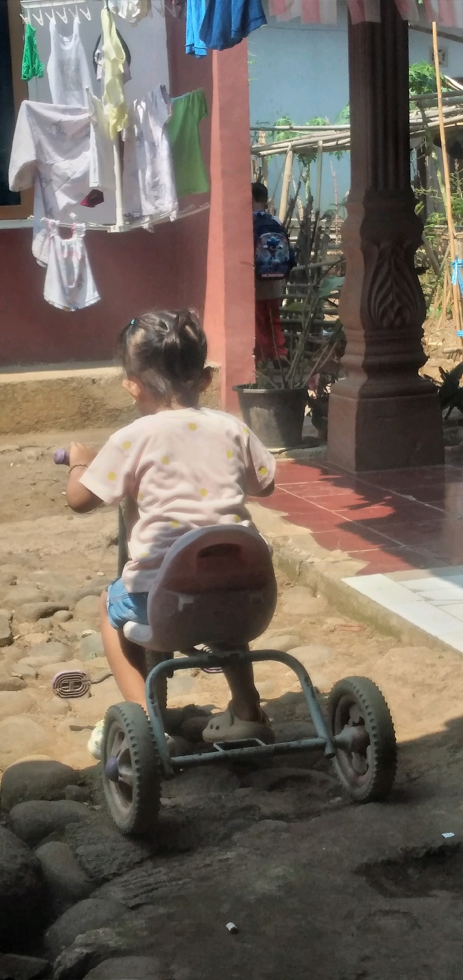 In Rahong, Indonesia, a young girl, viewed from behind, sits on a red tricycle. She wears a pink t-shirt adorned with yellow polka dots and denim shorts, and has her hair styled in two buns. The tricycle has a red seat and blue frame with three wheels. She is positioned on a rough, rocky surface. In the background, laundry hangs on a clothesline attached to a pink structure. To the right, a decorative dark brown pillar stands near potted plants and a fence made of bamboo. A person wearing a backpack with a cartoon character is visible further in the background. The ground is a mix of dirt, rocks, and red tiles.