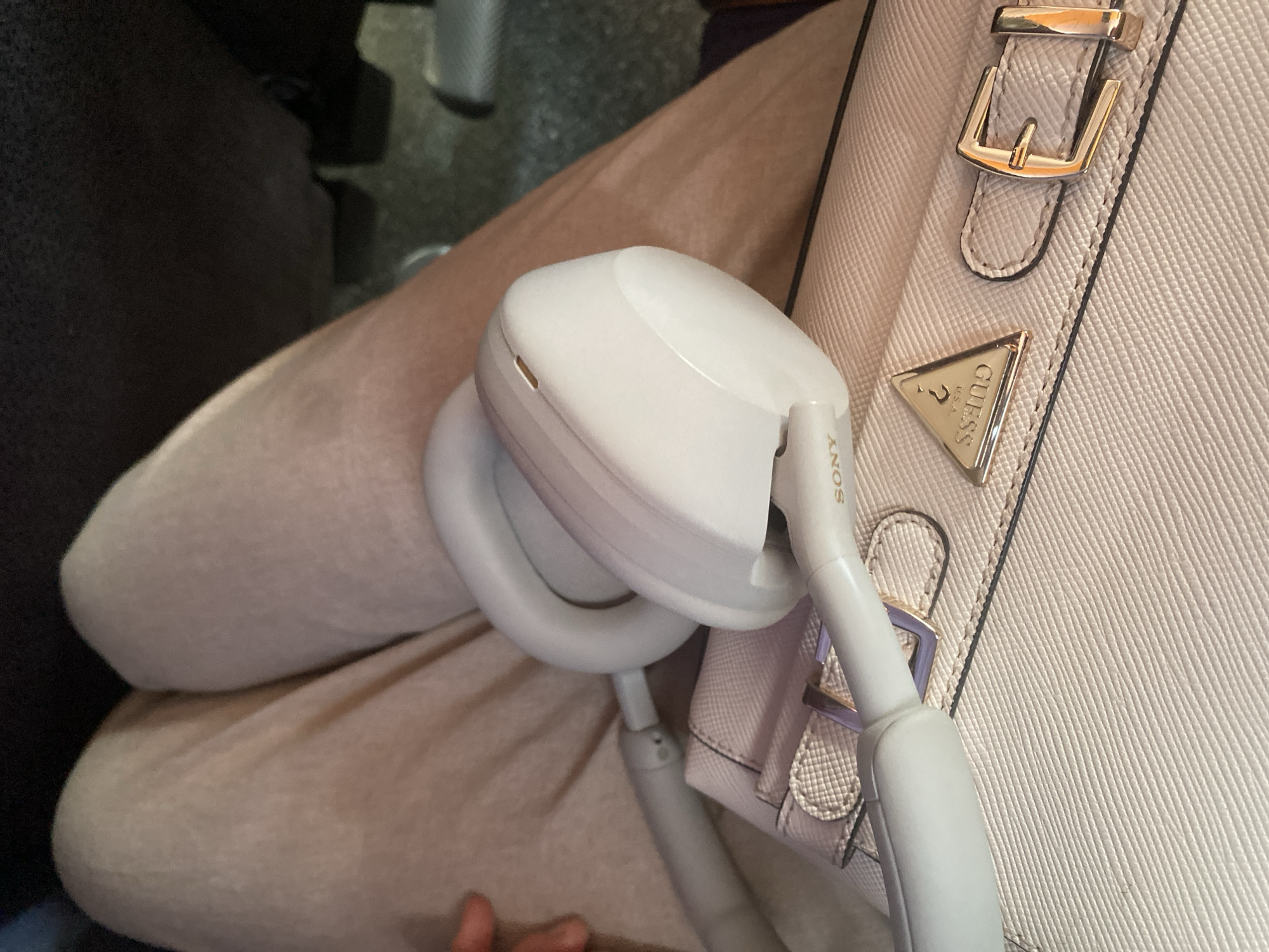 This media file is a close-up shot of a person's lap with a light-colored handbag and white headphones resting on it. The handbag is a light pink or beige color with a textured Saffiano finish. It features a gold buckle and a triangular metal emblem with the "GUESS USA" logo. The headphones appear to be wireless, with "SONY" branding visible on one of the earpieces.
The setting suggests a casual, indoor environment, possibly a car or a waiting area, given the seating visible in the background. The lighting is soft, implying it could be daytime or the light is artificial. The person's legs are covered in light-colored pants or leggings, with a hint of a darker garment underneath. There is no explicit indication of the time of day or weather. The image does not convey any strong emotions; it's a straightforward depiction of personal items. The text "SONY" and "GUESS USA" are clearly visible on the objects. No other significant details or activities are evident. The provided location context "Budva, Montenegro" does not offer any visual cues within the image itself.