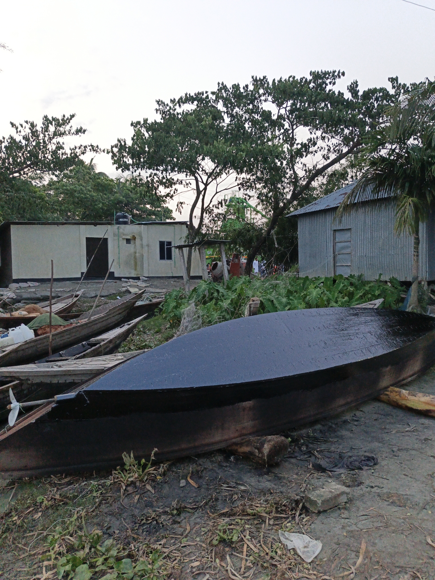 A landscape image shows a dark-colored boat in the foreground, lying on muddy ground with scattered vegetation. Behind the boat, a collection of other smaller wooden boats are visible. In the middle ground, there is a variety of green plants and foliage. Several buildings are present in the background, including a light-colored structure with a dark door and windows, and a corrugated metal building next to a palm tree. The sky is overcast, and the overall scene appears to be a rural or semi-rural location.