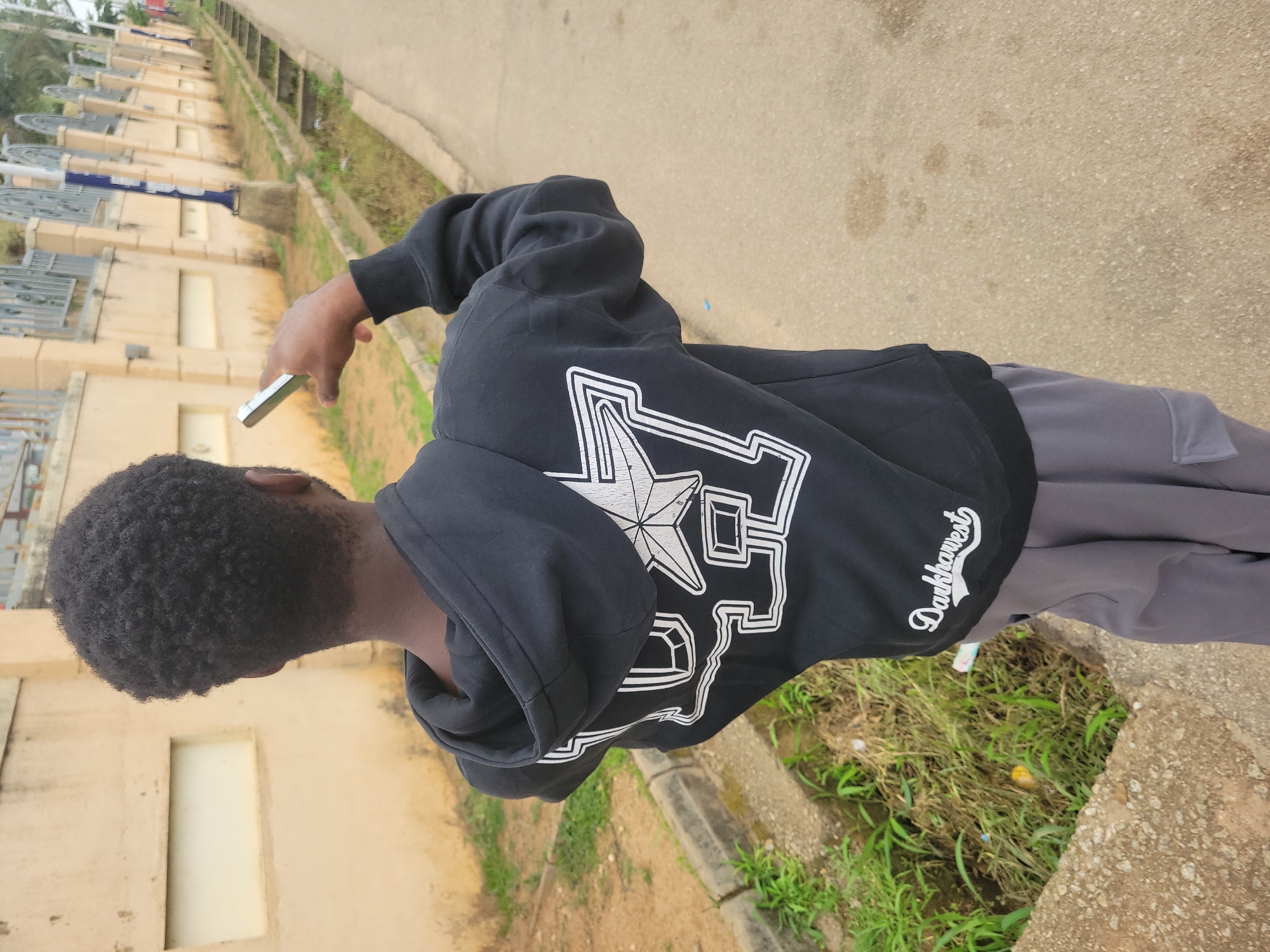 The image shows a person from behind, wearing a black hoodie with a white graphic design and grey pants. The person is holding a silver smartphone up, possibly to take a picture or record a video. The background suggests an outdoor urban environment. There is a tan-colored building with rectangular window-like indentations on the left. A drainage ditch with overgrown green grass and some debris runs along the right side of the paved path. A metal fence with decorative pillars runs along the top of the building. The overall impression is a street scene, possibly in a residential or institutional area. This scene could be located in Jos, Nigeria, given the architectural style of the building and the general environment.