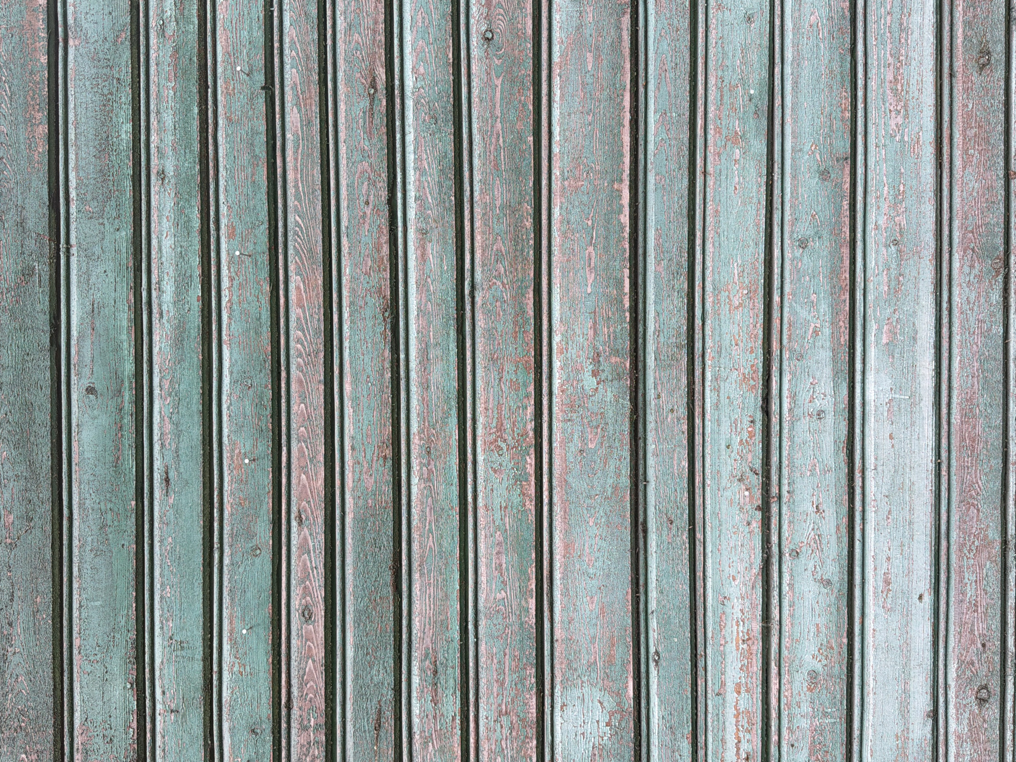 The image is a close-up, full-frame shot of a weathered wooden wall. The wall is constructed of horizontal planks with a faded, peeling teal-colored paint. The wood grain is visible beneath the paint, and there are areas where the paint has chipped away, revealing a reddish-brown underlayer. The planks are uniformly spaced, and some appear to have small nail heads or indentations. The lighting is even, suggesting an overcast day or diffused indoor lighting. There are no people or specific landmarks visible in the image, only the texture and surface of the wooden wall. The image is likely a detail shot intended for texture or material reference.