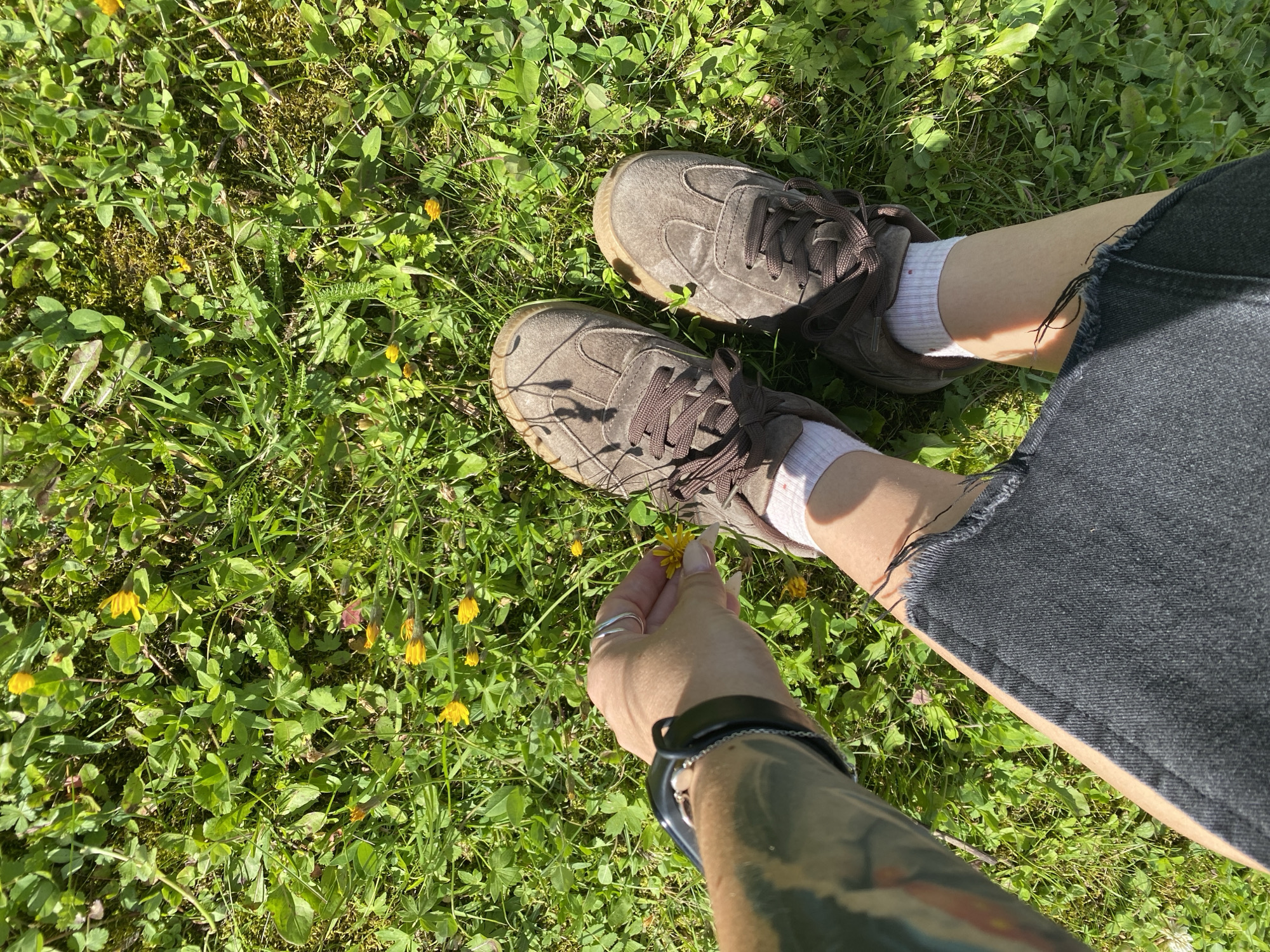 A person's legs, clad in dark denim shorts and brown lace-up sneakers with white socks, are visible standing in a grassy area dotted with small yellow flowers. The person's left hand, adorned with a ring and a smartwatch, is reaching down to pick one of the yellow flowers. The ground is covered in lush green grass and various small green plants. The scene is illuminated by bright sunlight, casting shadows on the grass and the person's shoes. The location is identified as the city of Foky, Russia.