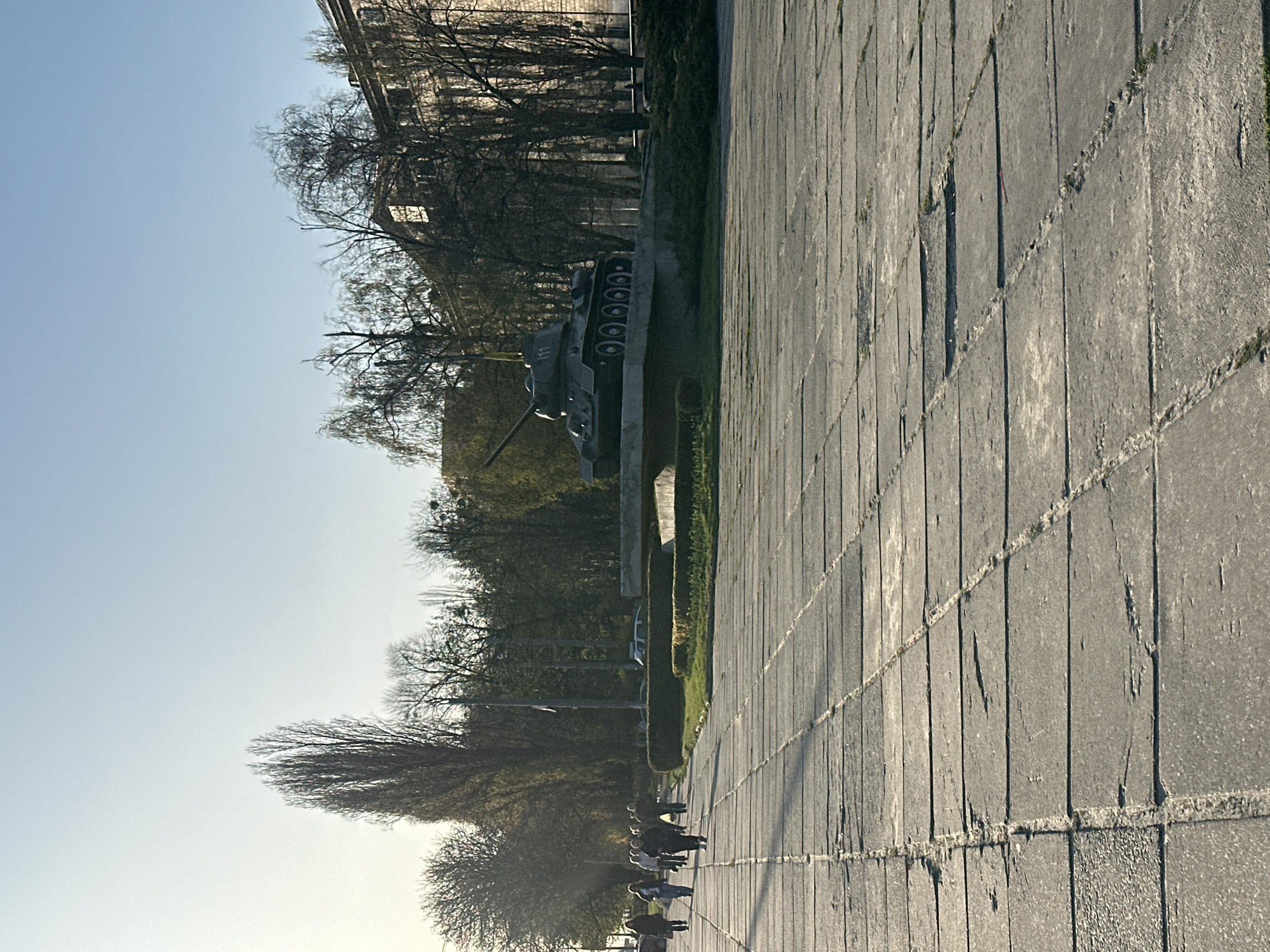 The image shows a tank monument in Kyiv, Ukraine, positioned on a concrete pedestal. To the right of the tank monument lies a paved area constructed from rectangular slabs, and a trimmed hedge borders the tank monument on the left. Several bare trees and a building can be seen in the background. In the foreground, a small group of people are walking along the edge of the paved area, away from the viewer. The sky is clear and light blue. The angle of the image suggests the picture was taken from a low vantage point.
