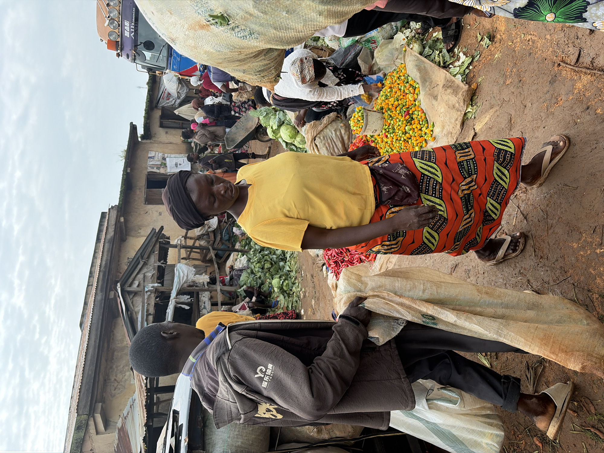 The image depicts a bustling outdoor market scene, likely in the city of Gura Top, Nigeria, with an overcast sky. In the foreground, a man wearing a dark jacket and dark pants carries a large, light-colored sack over his right shoulder and holds another sack in his left hand. Beside him, a woman in a yellow t-shirt and a vibrantly patterned orange and black skirt walks towards the viewer.
Behind them, the market is filled with people, various stalls, and goods. Piles of green leafy vegetables, including cabbage, are visible, along with a bucket filled with small, brightly colored peppers. A large truck is parked to the right, its front visible. The ground is dirt and appears somewhat muddy. Several people are engaged in activities like sorting produce or standing near stalls. The architecture of the market stalls suggests a rustic and traditional setup, with corrugated metal roofs and simple wooden structures. The overall atmosphere is one of a busy marketplace with active commerce.