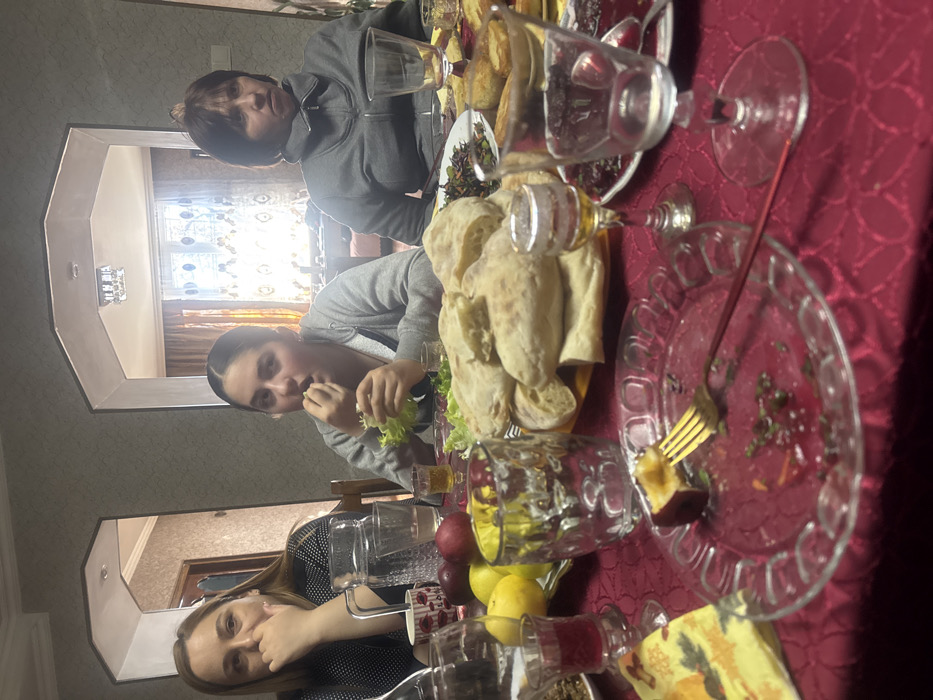 This image depicts an indoor dining scene within a residence in Bolnisi, Georgia, featuring three females seated around a table during what appears to be a meal.

On the left, a woman with long, light brown hair and a dark patterned top is visible, resting her left hand near her chin. In the center, a younger female with dark hair pulled back, wearing a grey hooded sweatshirt, is actively eating, bringing a piece of lettuce to her mouth with her right hand. On the right, a woman with short, dark hair, dressed in a grey zip-up jacket, maintains a neutral expression, looking forward.

The table is covered by a deep red, patterned tablecloth. In the immediate foreground, a clear glass plate with a fluted edge contains a partially consumed dark-skinned fruit, impaled by a gold-colored fork with a red handle, alongside scattered green fragments. Adjacent to it are a small stemmed glass containing an amber liquid, an empty clear stemmed glass, an empty clear textured glass, a small glass holding a dark red liquid, and a yellow patterned napkin.

Further back on the table, a large platter is stacked with numerous pieces of light-colored flatbread. To its left, a plate holds fresh green lettuce leaves, beside which another plate displays whole yellow and red fruits. A clear glass pitcher filled with water is also present. Other dishes with food, including a dark red preparation and golden-brown baked items, are partially visible in the background on the table.

The room features light grey/beige textured wallpaper. Two arched doorways in the background lead to adjoining rooms. The room visible through the right archway appears brighter due to a distant window, while the room accessed via the left archway is darker, revealing a dark wooden door.