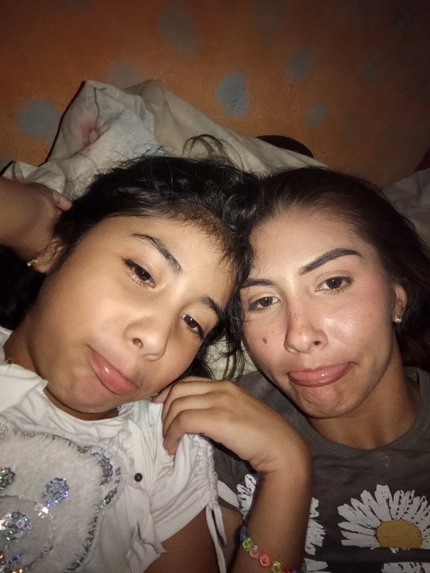 Two females are cheek to cheek in an indoor setting. The female on the left is younger and wearing a white shirt with a sequined pattern. She has dark hair. The female on the right has dark hair, darker skin, and is wearing a gray shirt with a flower design. Both are sticking their tongues out slightly. The backdrop is an orange wall and a white blanket. The location is Juan Griego, Venezuela.