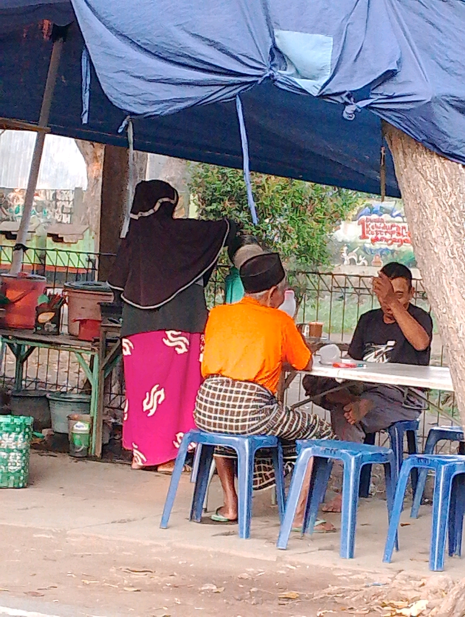 The image depicts a street scene under a blue tarp, likely an outdoor food stall or gathering area in Probolinggo, Indonesia. In the foreground, three individuals are seated or standing. A person wearing a black hijab and a vibrant pink skirt with white swirls is standing, facing away from the camera. To their right, a man in an orange t-shirt and a patterned sarong sits on a blue plastic stool. Another man, wearing a black t-shirt and dark pants, is seated opposite the man in orange, covering his face with his right hand. A white table is set up between the two seated men, with cups and other small objects on it. In the background, a metal fence separates the area from a wall adorned with a colorful mural. Various buckets and containers are visible on a makeshift shelf to the left of the frame, suggesting a functional setup for a business or community event. The ground is paved with concrete, showing signs of wear and tear, and scattered debris. A tree trunk is partially visible on the right side of the image. The overall atmosphere suggests a casual, everyday encounter in a tropical or subtropical climate.
