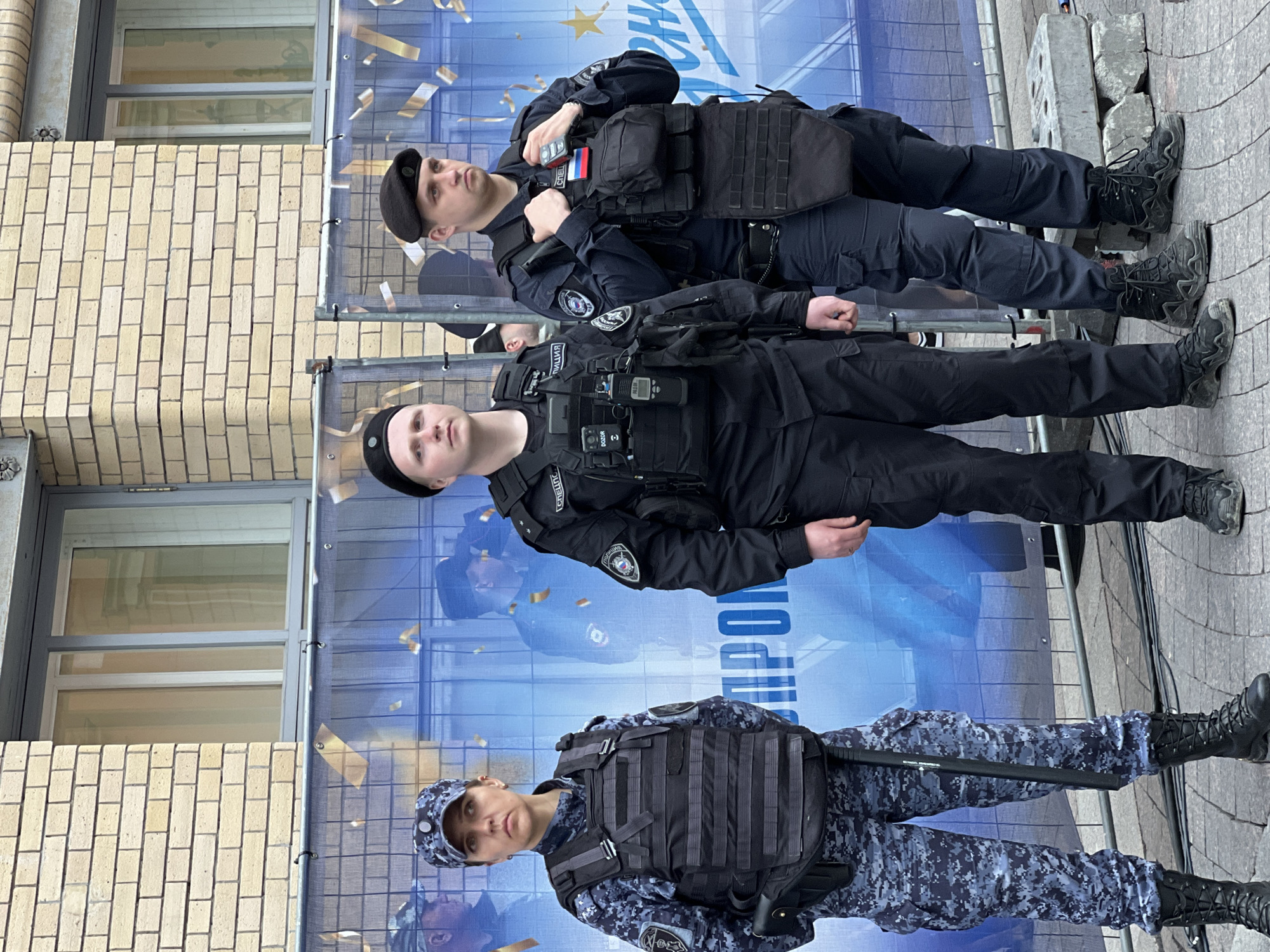 Three individuals, likely law enforcement personnel, stand in front of a blue backdrop featuring the words "ПОЛИЦИЯ СПЕЦНАЗ". The individuals are wearing dark tactical gear and headwear, with the first two from the top wearing berets and the bottom one wearing a camouflage cap. The top individual has a Russian flag patch. The person at the bottom is holding a baton. The location appears to be an urban area, possibly in Saint Petersburg, Russia. There is a building visible on the left side of the image with brick walls and windows. There is a metal fence present. The ground appears to be paved with grey stones.