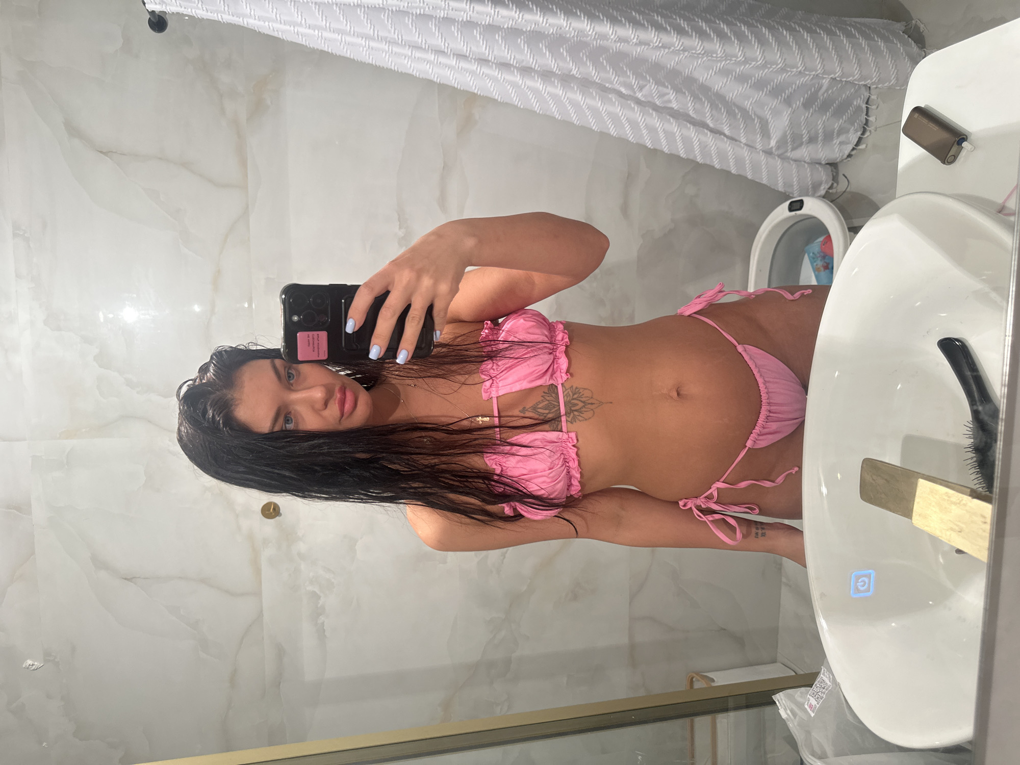 A woman in a pink bikini takes a selfie in a bathroom. The bathroom has a white marble wall, a white shower curtain, and a white sink. The woman is holding a black phone. The location of the image is a bathroom. The image contains no elements to identify it as Moscow, Russia.