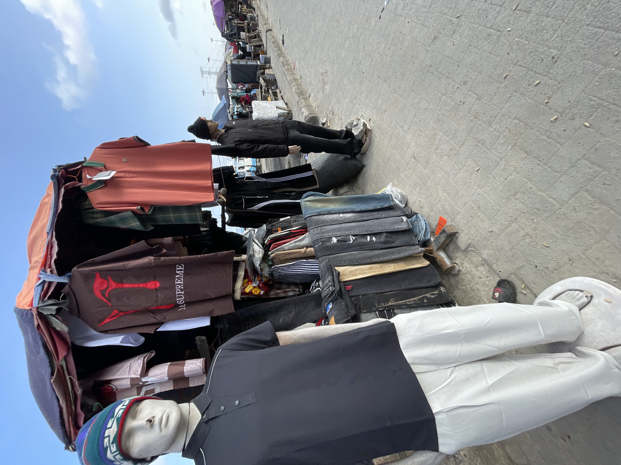 The image depicts an outdoor clothing stall located in the city of Lekki, Nigeria, under a clear sky with scattered clouds. The stall features an assortment of men's apparel, including polo shirts, t-shirts, and trousers, displayed on mannequins and hangers. A mannequin on the left is dressed in a black polo shirt and light-colored trousers, while another on the right wears a black jacket and dark trousers. Several other clothing items, such as jeans and striped shirts, are visible hanging or folded. In the background, the scene extends to a busy street with more stalls, people, and vehicles, suggesting a market environment. The ground is paved with interlocking bricks.