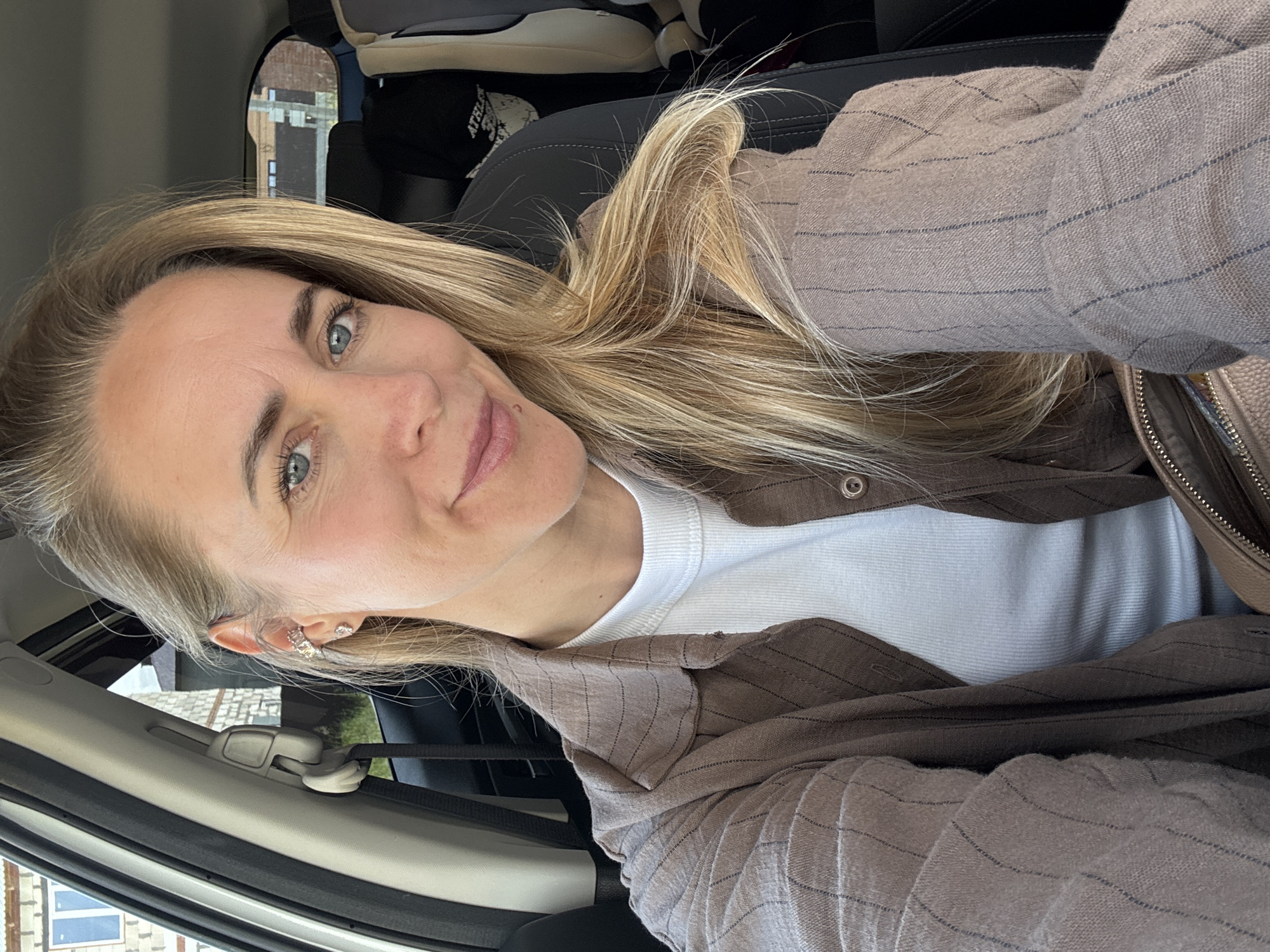 A woman with blonde hair and blue eyes is taking a selfie in the driver's seat of a car. She is wearing a white ribbed tank top and a brown pinstripe open button-up shirt. Her left ear has a silver cuff earring. The background shows a car interior with a seatbelt and a car seat, and through the window, buildings and greenery are visible. The woman appears to be in the city of Chaikovsky, Russia, based on the architectural style of the buildings.