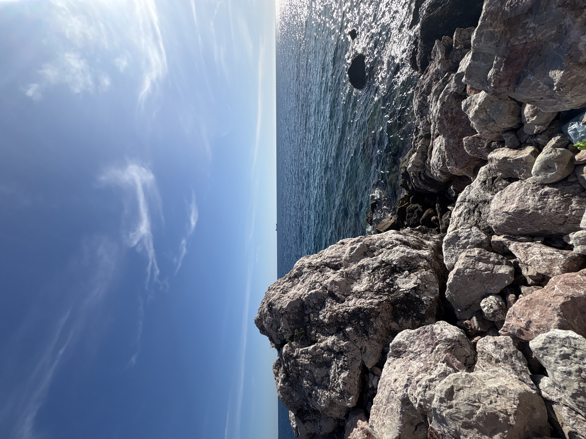 The image shows a rocky shoreline in Bar, Montenegro. The focus is on large, light-colored rocks that make up the immediate foreground. To the left of the rocks is a view of the blue sky with wispy clouds. To the right of the rocks, the Adriatic Sea is visible, shimmering with sunlight. The horizon line is clear and separates the sea from the sky.