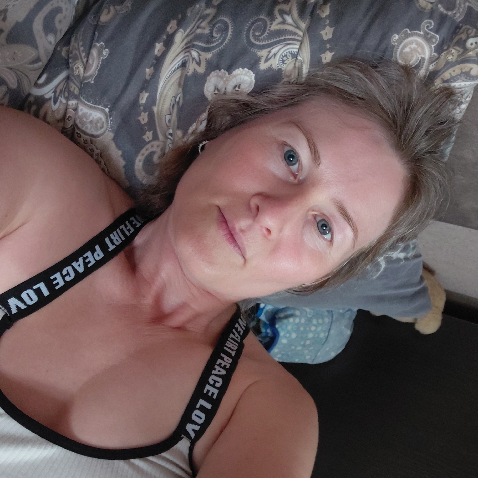 A woman with graying blonde hair and blue eyes is lying on her side on a patterned pillow and appears to be taking a selfie. She is wearing a white tank top with a black band that has the words "LOVE FLIRT PEACE LOVE" printed on it. The background shows a dark surface, possibly a sofa or bed, and a pillow with a light blue and white pattern. The scene is indoors, and there are no explicit indicators of location within Novosibirsk, Russia.