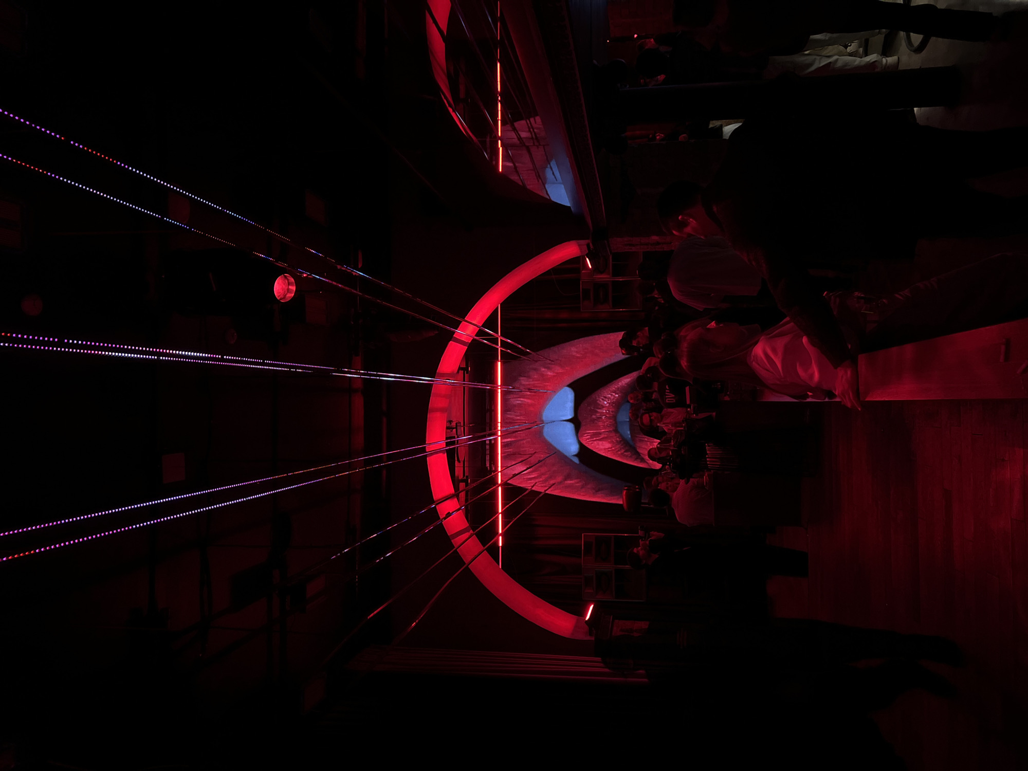 The media file displays a dimly lit indoor scene, dominated by artificial light, set in what appears to be a modern venue such as a club, bar, or art installation in Vladivostok, Russia.
On the left side of the image, multiple parallel lines of small, glowing LED-like dots, in shades of pink, purple, white, and blue, stretch diagonally across a dark background. A single, distinct red glowing orb is also visible in this dark space.
The central part of the image features a large, prominent, semi-circular arch glowing vibrantly with red light. Inside this red arch, a wavy, textured surface emits a contrasting bright blue light. Several thin, vertical red light strips are reflected within this blue area. Further back, another similar, though less distinct, red-lit arch with a blue interior can be seen, suggesting a repeating architectural or decorative element.
On the right side of the image, several people are gathered, mostly in silhouette or partially illuminated by the red light. One person in the foreground wears a white top and is looking downwards. Their features are indistinct due to the low light. The floor beneath them appears to be a reddish-brown, possibly wood, reflecting the ambient red light.
The time of day is clearly night, or the scene is entirely indoors without natural light. The strong red and blue lighting creates a moody, futuristic, and dramatic atmosphere. Faint white text, possibly "DVI" or "DV1," is visible on a dark object in the upper right background, near the group of people.