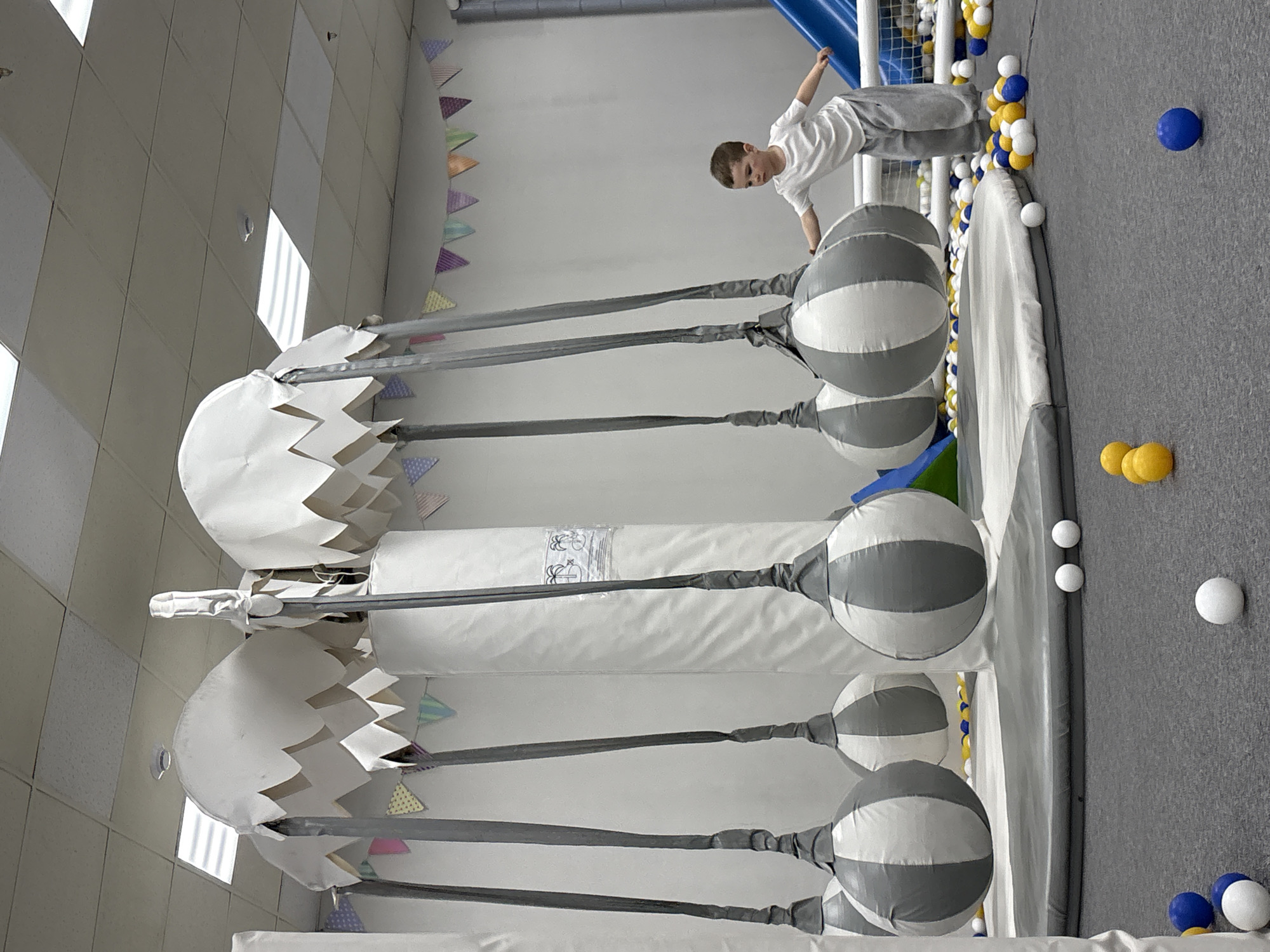 The image shows a child in an indoor playground. The location appears to be in Krasnoyarsk, Russia, based on visual clues. The child is wearing a white t-shirt and grey pants, leaning over a structure with grey and white striped ball-shaped objects attached to poles. Adjacent to this is a ball pit with blue, yellow, and white balls scattered around. The walls are white, and there's a decorative banner with small triangle flags along the top. A soft climbing structure is also visible.