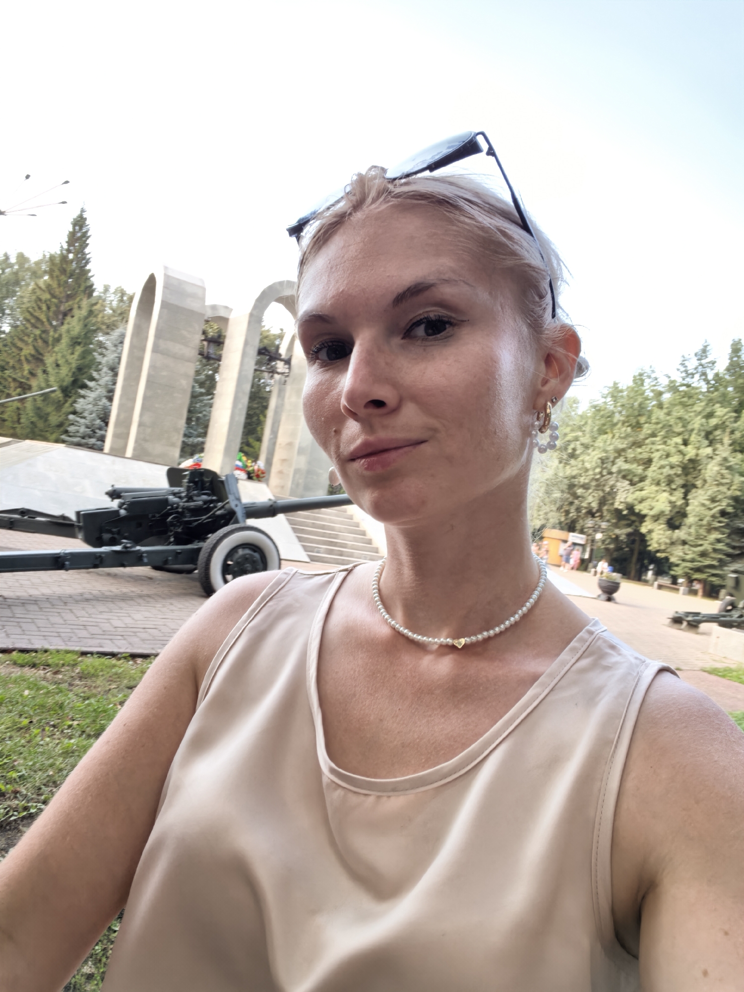 A young woman with blonde hair pulled back and sunglasses perched on her head takes a selfie outdoors. She is wearing a light-colored, sleeveless top and a pearl necklace with a small gold heart pendant. She is also wearing hoop earrings adorned with pearls.
In the background, there is a war memorial featuring a large archway and what appears to be a cannon on display. Trees and a paved walkway are also visible. The scene suggests a historical or memorial site within a city, possibly in Sterlitamak, Russia, given the context of the prompt. The lighting indicates it is daytime.