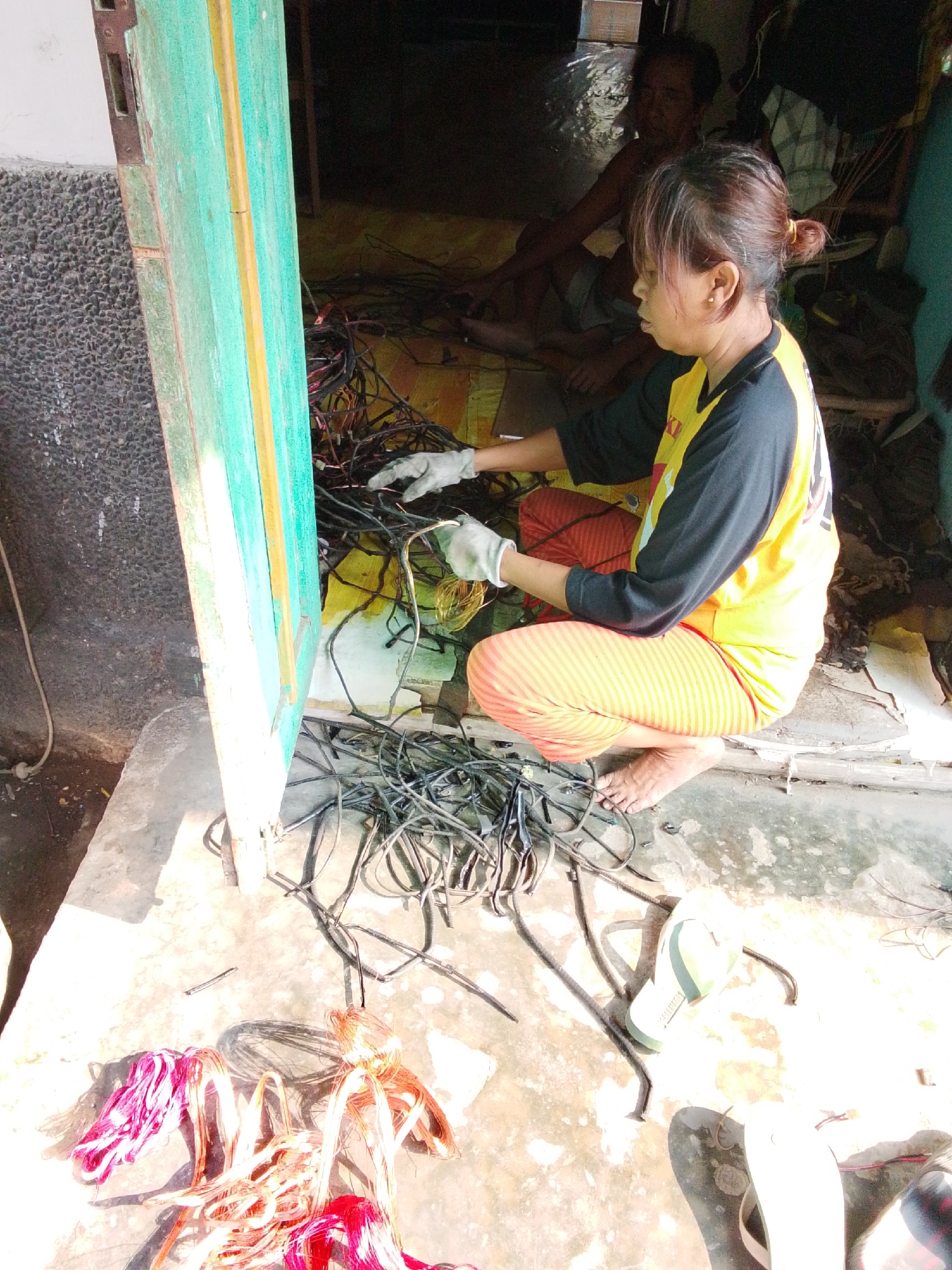 In Bulurejo, Indonesia, a woman wearing gloves and a yellow and black shirt crouches on a concrete floor, sorting through a large pile of electrical wires and cables. She is focused on her task, with a man sitting in the background, partially obscured. The scene appears to be an outdoor workshop or storage area, with a dilapidated wooden doorway and a textured wall to the left. Several bundles of colorful wires are visible in the foreground, suggesting an ongoing project or salvage operation. The sunlight casts strong shadows, highlighting the rustic environment.