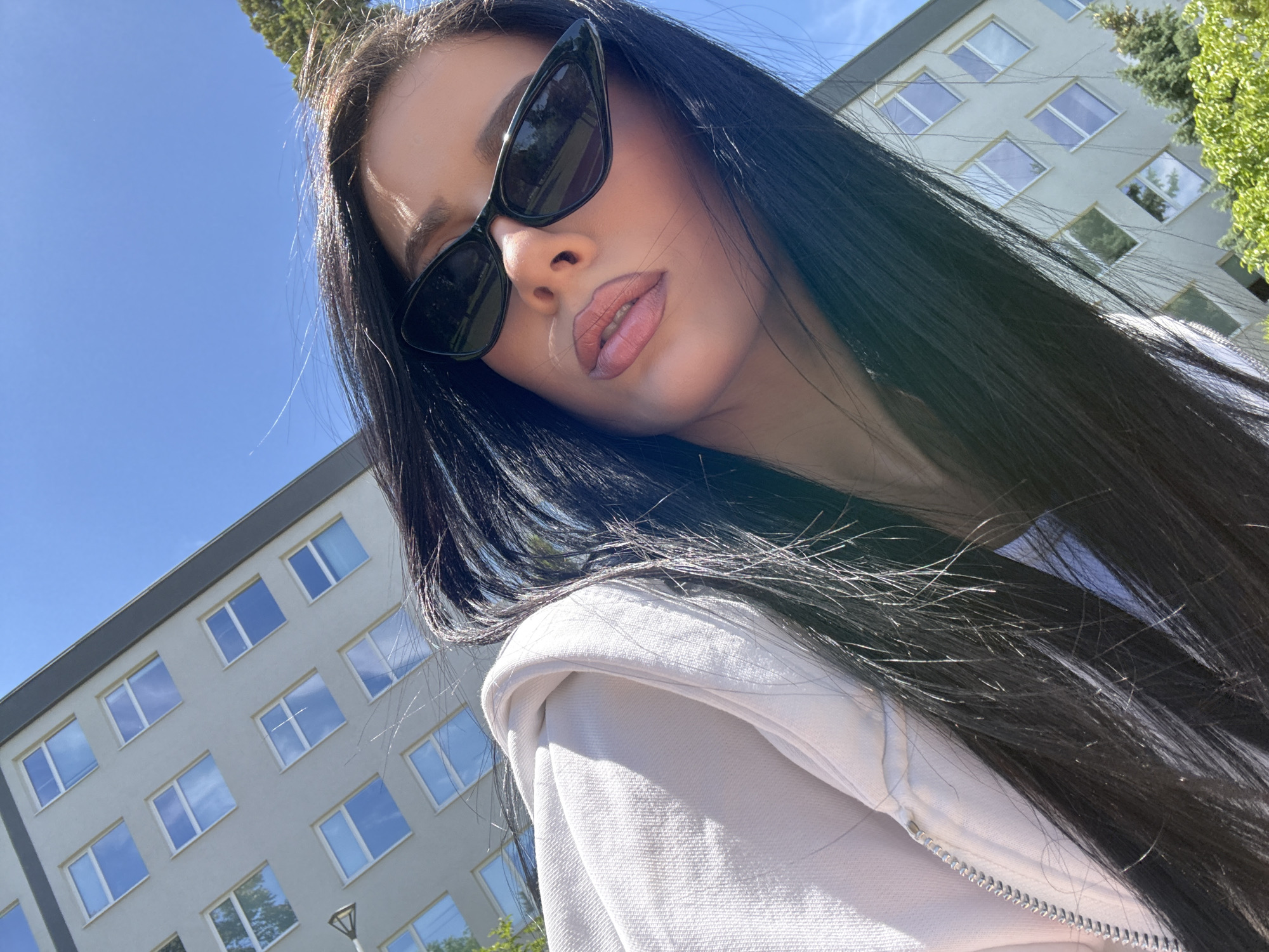 A low-angle, close-up shot shows a woman with long, dark hair and sunglasses outdoors. She's wearing a white jacket or hoodie. Behind her, there is a multi-story building with a grey facade and numerous square windows. The sky is a clear, bright blue. The exact location in the city of Unknown, Unknown cannot be determined from the image.