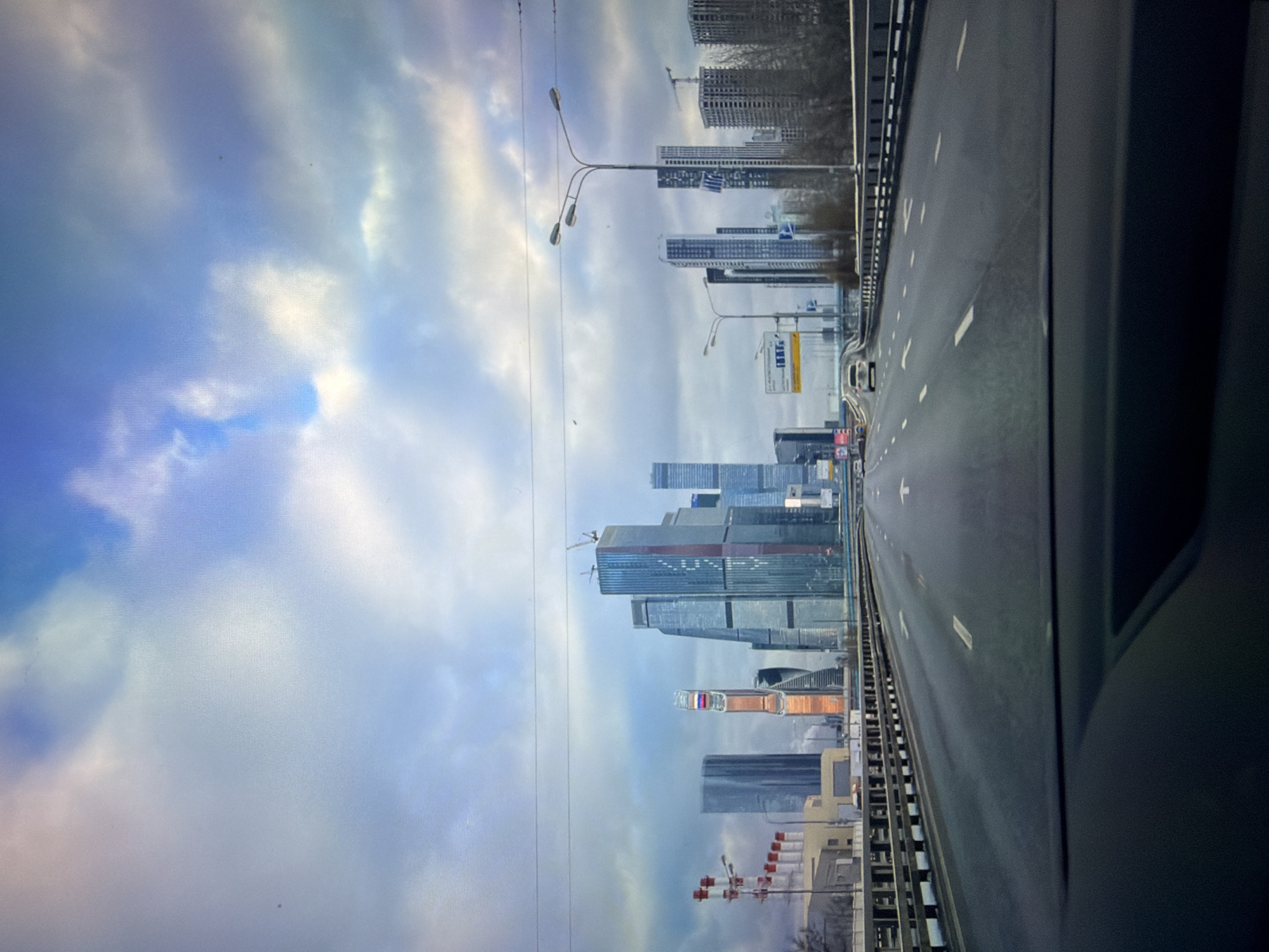 The image depicts a view from a vehicle traveling on a multi-lane highway towards a modern city skyline. The sky is cloudy with patches of blue. Tall, contemporary buildings dominate the background, some featuring glass facades and distinctive architectural designs. In the foreground, the highway stretches forward with clear lane markings and a few vehicles visible. A guardrail lines the right side of the road. The overall scene suggests a metropolitan area, likely during daytime, given the lighting conditions. The location is specified as the city of Kotelniki, Russia.
