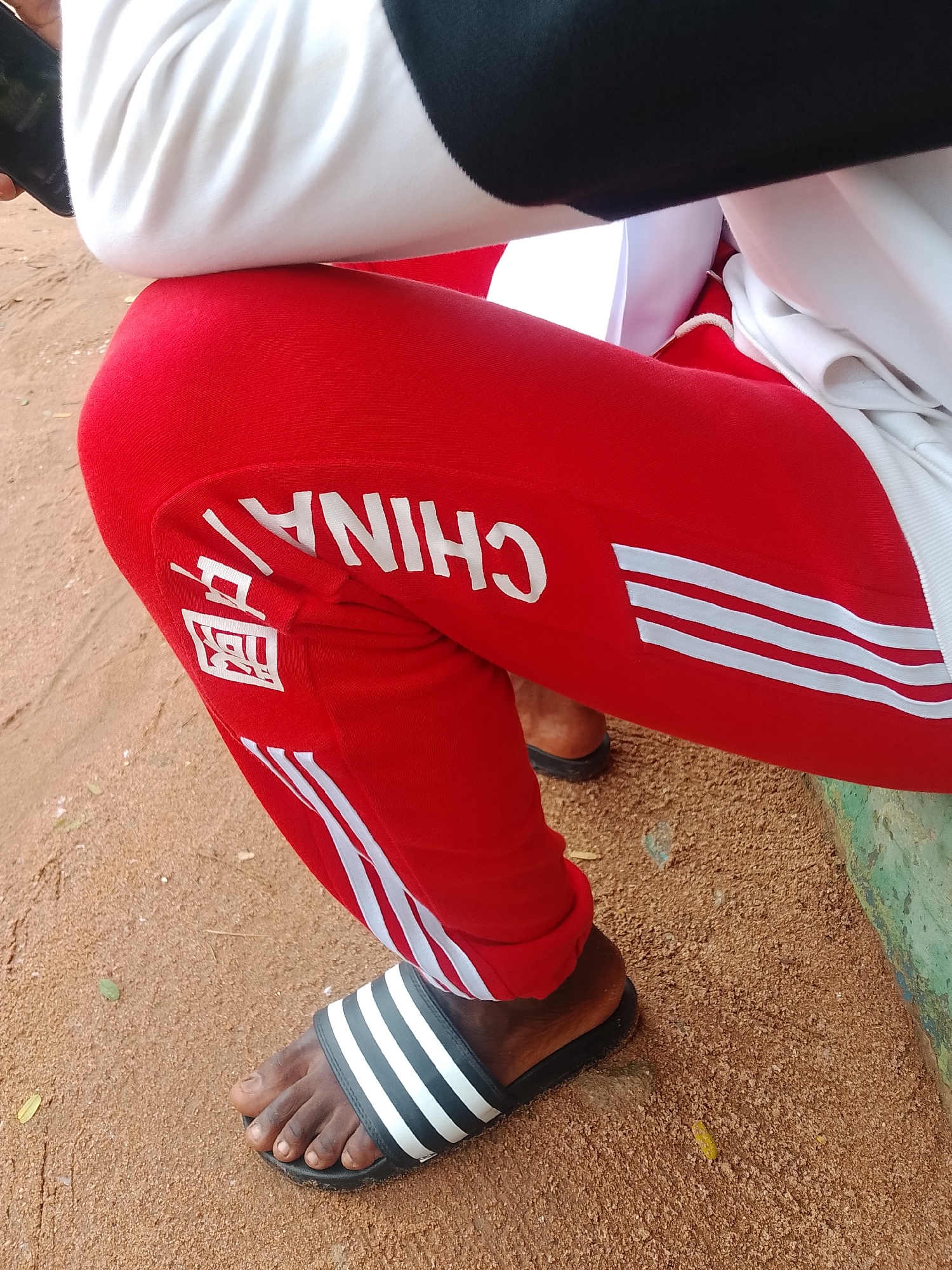 A close-up, eye-level shot shows the lower half of a person wearing red sweatpants with white lettering and stripes on the side. The lettering, in white, appears to be in English and Chinese characters. The person is wearing black and white Adidas slide sandals. They are sitting on a bench, with their legs crossed, in an outdoor, sandy environment. The background consists of sand and a green object, possibly a bench or a wall. The lighting is natural, suggesting daytime. This scene is consistent with informal public spaces in Jalingo, Nigeria.