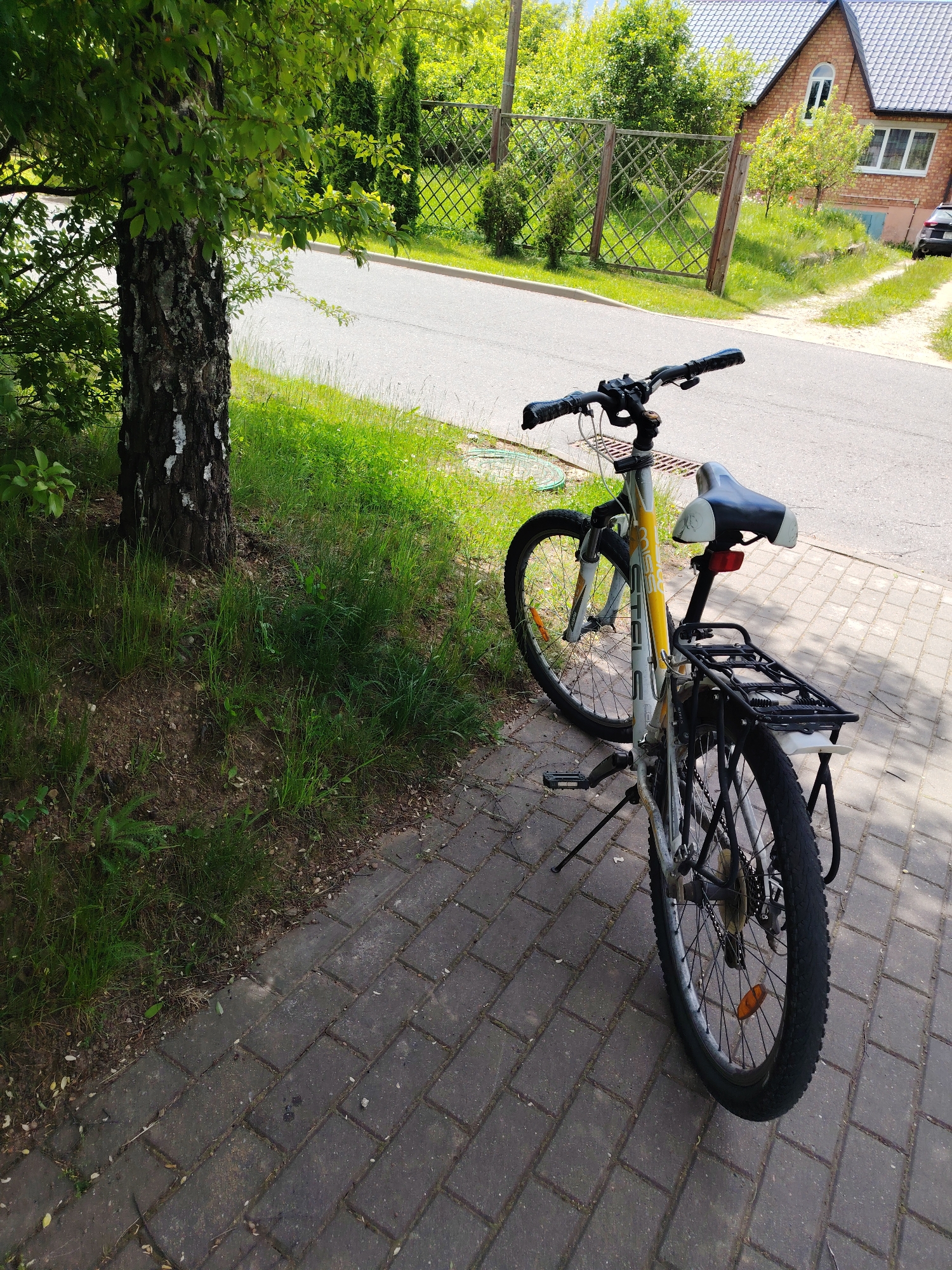 The image displays an outdoor scene on a clear, sunny day in what appears to be a residential area within Бараўлянскі сельскі Савет, Belarus. In the foreground, a mountain bicycle is parked upright on a dark grey paved sidewalk. The bike has a silver frame with yellow accents and the brand name "STELS" partially visible. It features a black and white seat, a rear cargo rack, and a red reflector.
To the left of the bicycle, a tree with distinctive white and dark bark, possibly a birch, stands tall with vibrant green leaves. Green grass grows around the tree's base and along the edge of the sidewalk, which borders an asphalt road. The strong sunlight casts prominent shadows from the tree and the bicycle, indicating it is likely midday or early afternoon.
In the mid-ground, the asphalt road curves slightly. Beyond it, a sturdy wooden fence with a diagonal lattice design stretches across the scene. Behind the fence, several green bushes and trees contribute to the lush, verdant surroundings. On the right side of the background, a two-story brick house with a dark tiled roof and multiple windows is visible. A dark car is partially seen parked near the house's driveway. No people are present in the image.