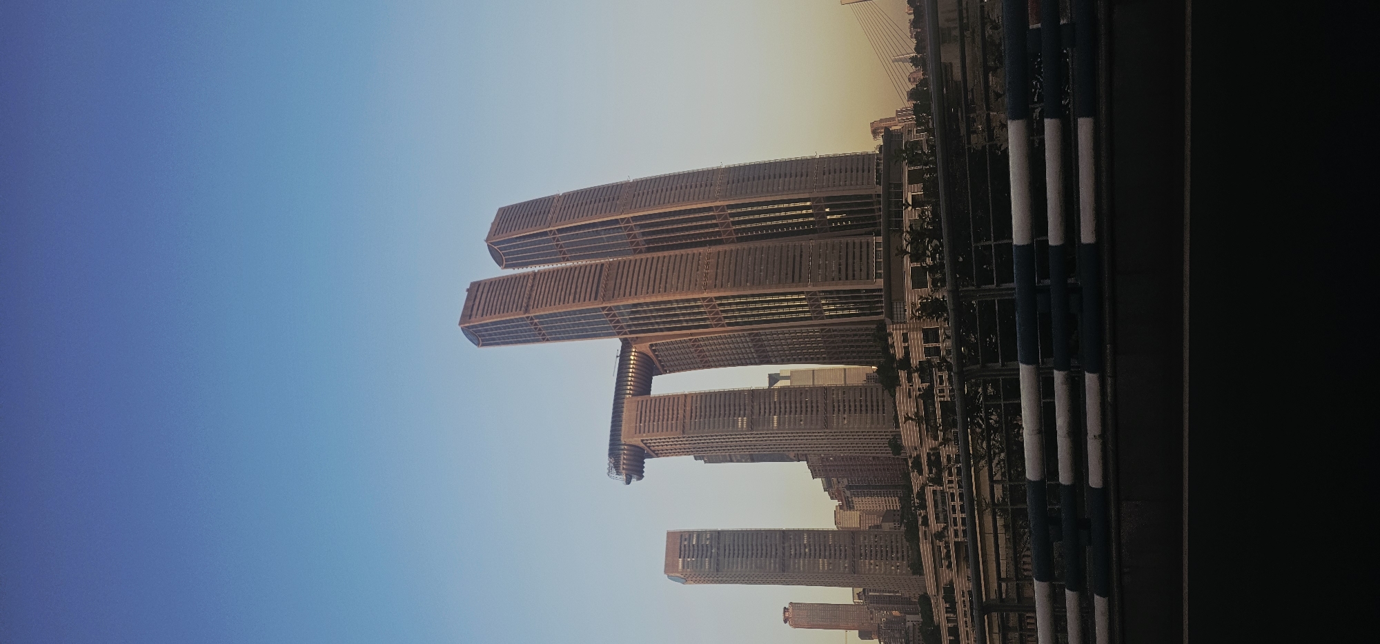 The image depicts the Raffles City Chongqing complex in the Nan'an District of Chongqing, China, during what appears to be either sunrise or sunset. The sky is a clear gradient of light blue to pale yellow, indicating a low sun angle. The primary subjects are the distinctive, interconnected skyscrapers of Raffles City, with their unique curved design and elevated sky bridge. Several other modern buildings, varying in height and architectural style, form a dense urban skyline behind the Raffles City complex. In the foreground, a dark, possibly asphalt surface runs horizontally across the bottom of the frame. Above this, a barrier made of dark metal fencing with alternating white and dark blue horizontal bars is visible. Behind the barrier, sparse greenery and the lower levels of buildings are partially obscured. No people are visible in the frame. The overall impression is of a contemporary cityscape captured during the transitional light of day.