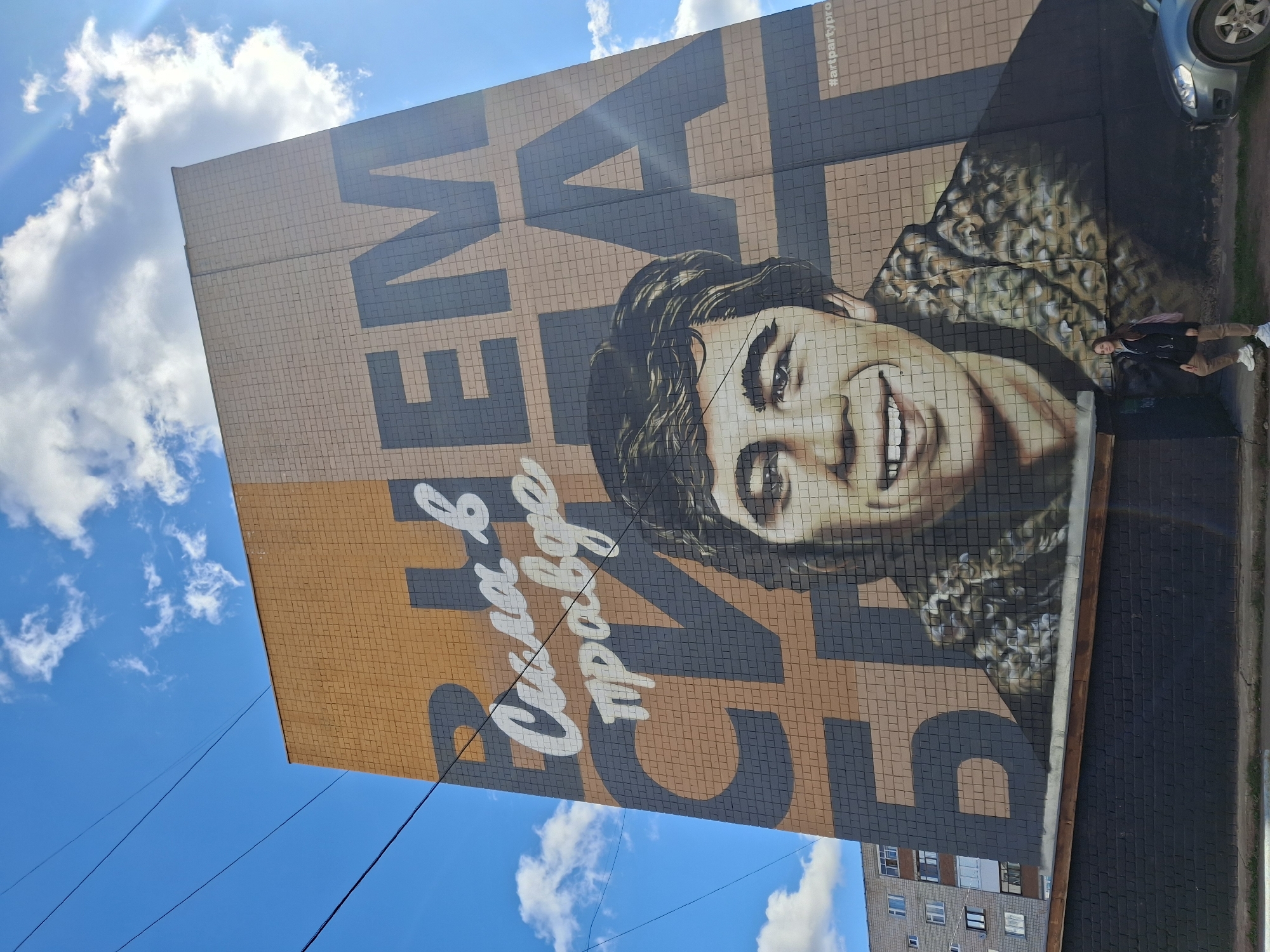 This media file captures a large mural painted on the side of a building in Sarapul, Russia, viewed from a slight upward angle under a bright blue sky with scattered white clouds. The mural, rendered in a mosaic-like style on a tiled or brick wall, features a prominent portrait of a smiling young person with dark, wavy hair, looking upwards and to the viewer's left.
To the left of the portrait, large Cyrillic text is painted in white against a dark gray background, stating: "В ЧЕМ СИЛА?" (In what is the power?). Below this, also in white against an orange-brown background, is the answer: "В ПРАВДЕ." (In the truth.). This is a famous quote from the popular Russian film "Brother." In the upper right corner of the mural, the smaller hashtag "#artpartypro" is visible.
A female individual stands on the sidewalk to the right of the mural, facing the camera and appearing to pose or observe the artwork. She is dressed in a dark jacket, light brown pants, and white sneakers. Behind her, a silver car is partially visible, parked on the street. The bright sunlight casts distinct shadows, suggesting it is daytime, likely morning or afternoon. Utility lines stretch across the clear sky in the lower left, and another residential building is visible in the distance on the lower right. The scene conveys a sense of a sunny day with public art on display.
