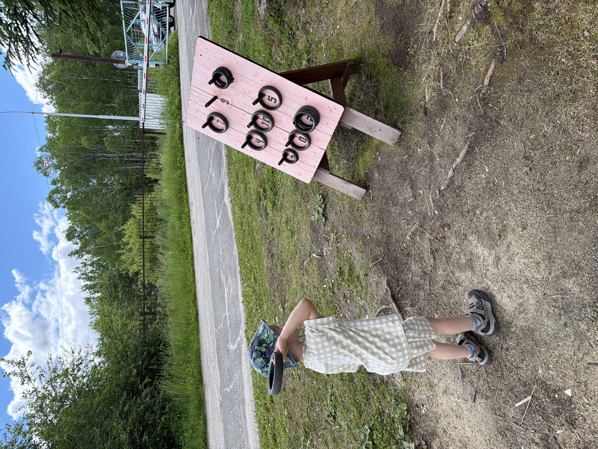 In Kul'dur, Russia, a young child stands on a gravelly and grassy area, holding a black ring in their right hand, preparing to throw it at a pink wooden board. The board, propped up on wooden legs, has pegs with rings of various sizes on them, each marked with a number from 4 to 10. The child is wearing a checkered shirt and shorts, a patterned bucket hat, and athletic shoes. Behind the child and the game, a paved path runs alongside a grassy area. Further in the background, a rusty metal fence, a white post, and a blue and white barrier gate are visible. Trees and dense foliage form the backdrop, with a bright, cloudy sky above.