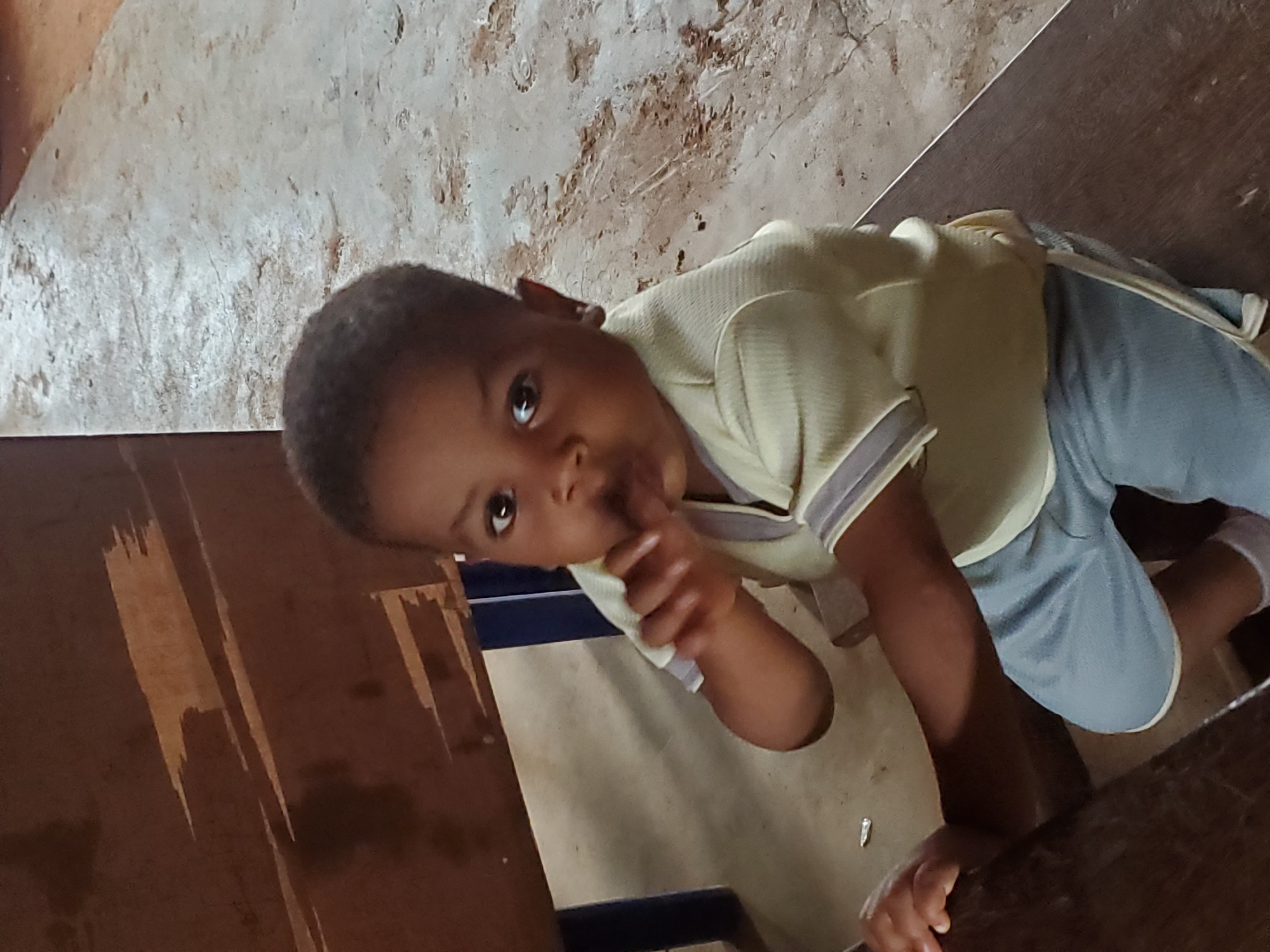 A child with dark hair and skin, wearing a light yellow shirt with gray trim and blue shorts, is kneeling on what appears to be a floor covered with a light-colored material or rug. The child is looking towards the camera with their right hand near their mouth. The background shows peeling paint on a wall and a wooden surface. The location is presumed to be Ukwu-Abwa, Nigeria.