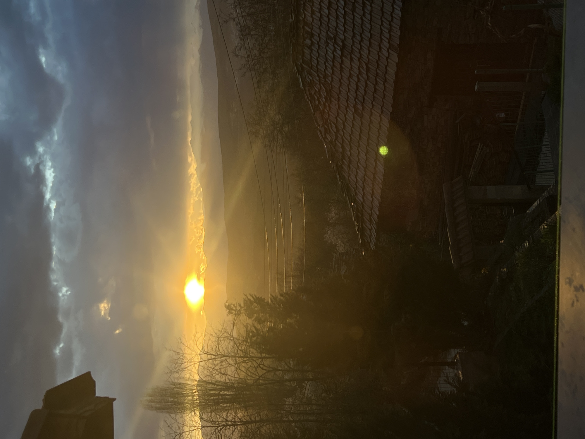 The image shows a landscape view in Kribul, Bulgaria, dominated by the setting sun. The sun is positioned low on the horizon, partially obscured by clouds, creating a bright, golden light that permeates the sky and surrounding environment. Utility poles and wires are visible extending across the landscape. In the foreground, silhouetted trees and buildings are present. To the right, a portion of a roof, covered with tiles is visible. The clouds range from dark grey to lighter hues, reflecting the sunlight. The time of day is most likely at sunset or dawn.