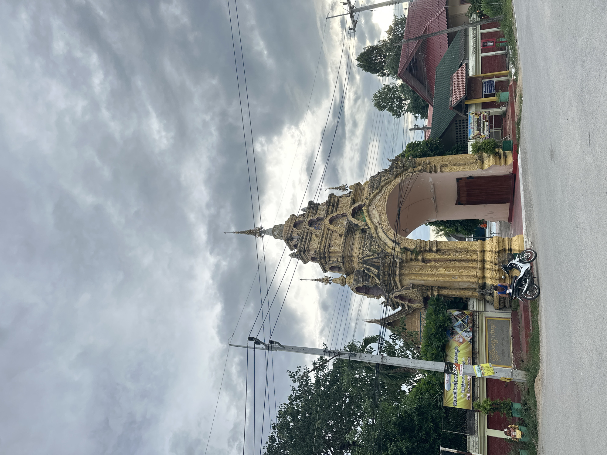 The image shows a street scene in Ban Patan, Thailand. A highly ornate golden archway, likely part of a temple, dominates the left side of the frame. The arch is intricately carved with spires and decorative elements, suggesting a traditional architectural style. A paved road runs along the right side of the image.
In front of the arch, there's a man sitting on a motorcycle, which is parked on the side of the road. Behind him, a building with a red roof and a closed wooden door is visible. Several trees and electrical wires add to the urban landscape. The sky is overcast with dramatic, cloudy formations, suggesting either early morning or late afternoon, or possibly impending rain.
Several signs are displayed on the roadside and on the building. One prominent sign, with Thai script, appears to announce an event or festival. The visible text includes "พิธีงานท", "ประเพณีสรงน้ำพระ 599 นวิสาขบูชา", and "วัดลัฏฐิวนารา". This suggests a religious event related to Songkran, Vesak, or a temple anniversary. The overall atmosphere is one of a typical day in a Thai town, with elements of daily life and religious significance present.