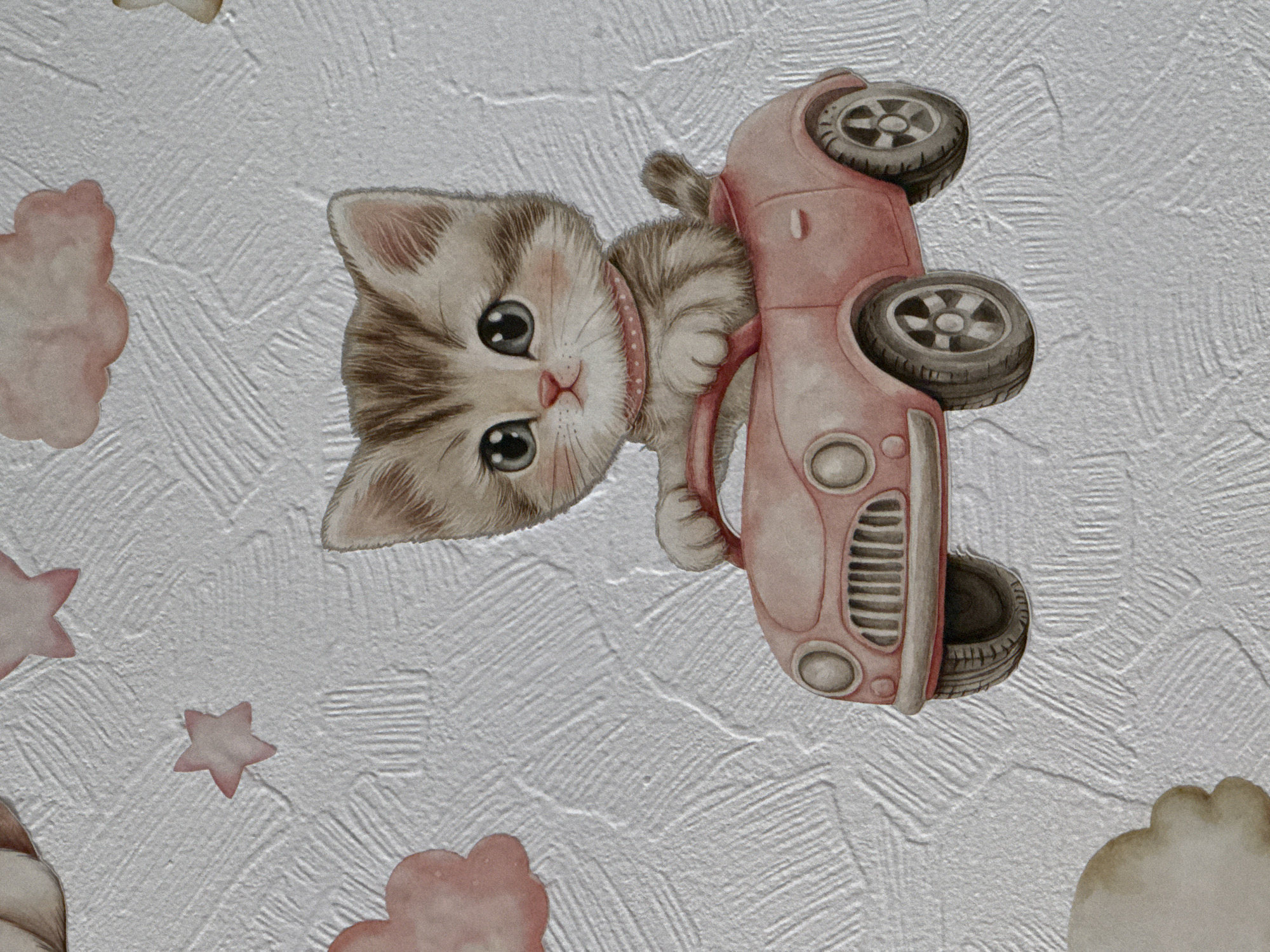 The image is a close-up of a textured white surface, possibly wallpaper, featuring cartoon-style illustrations. Dominating the scene is a drawing of a kitten holding onto a pink car. The kitten is light brown and grey with large, expressive eyes and is wearing a pink collar with white polka dots. The car is facing downwards, with visible wheels and a front grill. Several other cartoon elements are scattered around the image, including light pink clouds and stars. The location is not discernible from the image.