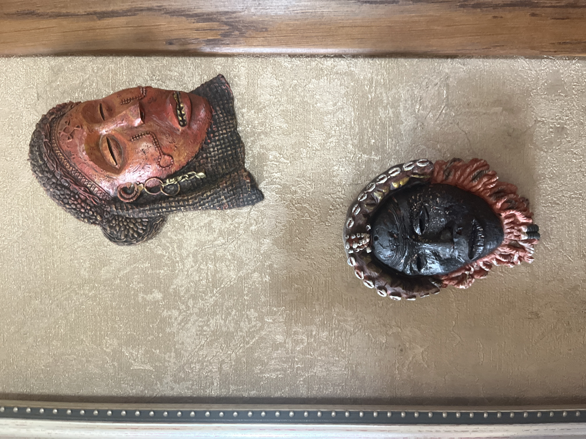 The image displays two African masks mounted on a textured, beige wall within a room. The wall has a slightly rough, embossed pattern. A framed border with a light-colored matte finish and a darker, studded inner edge is visible on the left side of the image. To the right, a portion of a wooden frame or furniture is present. The top mask is reddish-orange with intricate carvings and adornments, including what appears to be a fabric headdress and earrings. The bottom mask is black with a darker texture, adorned with shells around the headdress and a fringe of red and white threads around the face. There are no people or discernible actions depicted in the image. The location is specified as Vladimirovka, Russia, but the visual elements do not provide specific contextual clues to confirm or deny this.