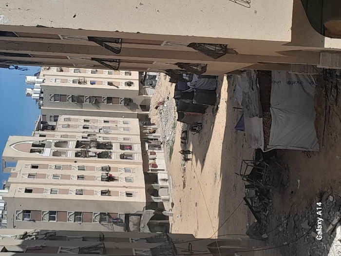 This image captures a wide-angle view of a heavily damaged residential area in Khan Yunis, Palestinian Territory, under clear daylight conditions. Multiple multi-story apartment buildings, predominantly beige with decorative architectural elements, stand in the background and midground. These structures display extensive damage, including numerous shattered and missing window panes, exposed interiors, partial balcony collapses, and visible facade deterioration with pockmarks and missing sections. Debris, rubble, and twisted metal frames are scattered across the unpaved, sandy ground in the foreground and midground. A cluster of at least five makeshift shelters, constructed from tarps, fabric, and corrugated materials, occupies the midground; one visible white tarp bears the "unicef" logo. Two individuals, dressed in dark clothing, walk on a dirt path in the midground, moving away from the viewer towards the damaged buildings. Several other indistinct figures are present closer to the buildings' bases. The foreground contains additional debris, including a large metal frame and exposed wires.