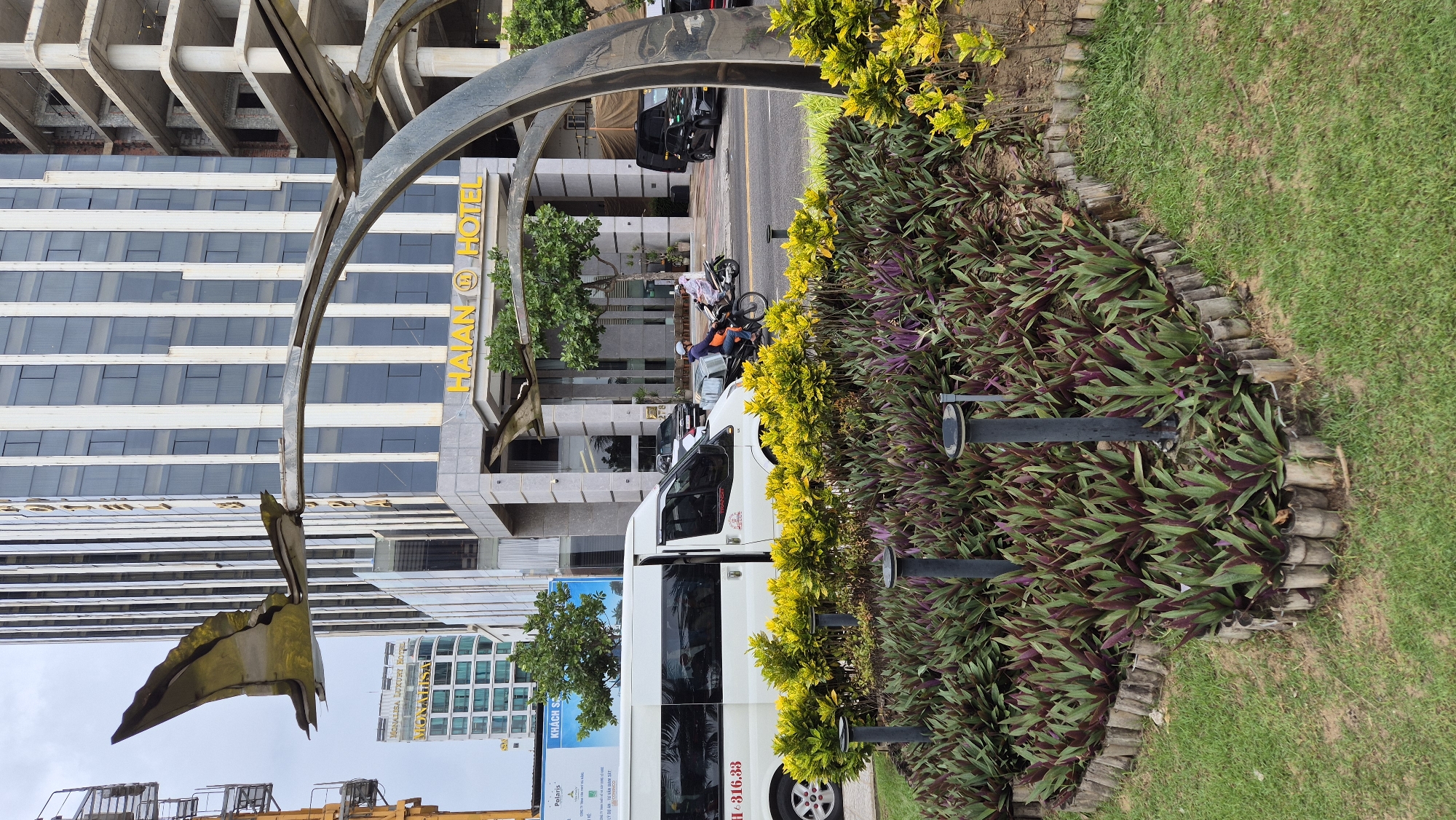 This image captures a street scene in Đà Nẵng, Vietnam, featuring the Haian Hotel prominently. In the foreground, a landscaped garden with bamboo edging contains low-lying purple and green plants and yellow flowers, interspersed with dark grey lampposts. A white van with open doors is parked on the street to the left, with partial lettering visible on its side including "H - 316.33". To the right of the van, a man on a motorcycle wearing a white helmet and a light-colored garment rides away from the camera, followed by another person on a scooter. The Haian Hotel's facade is visible behind the street, characterized by a modern glass and concrete design with large windows and yellow lettering above the entrance. To the left of the Haian Hotel, a taller building with a similar architectural style, identified as "MONALISA LUXURY HOTEL" on a sign, is also visible. A large, abstract metal sculpture resembling a bird in flight arches over the scene in the foreground. The sky is overcast. Construction is evident on the right side of the frame, with unfinished concrete structures of a building under development.