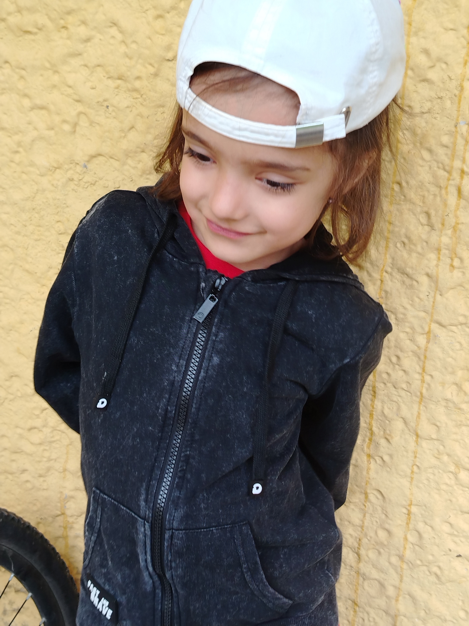 A young girl with brown hair, wearing a white baseball cap and a black hooded jacket over a red shirt, stands against a textured yellow wall. Her head is tilted down and to her left, with a slight smile on her face. The lower left corner of the image shows a portion of a bicycle wheel. The scene appears to be outdoors in a city setting. No specific location within Noviy, Russia, can be identified from the image.