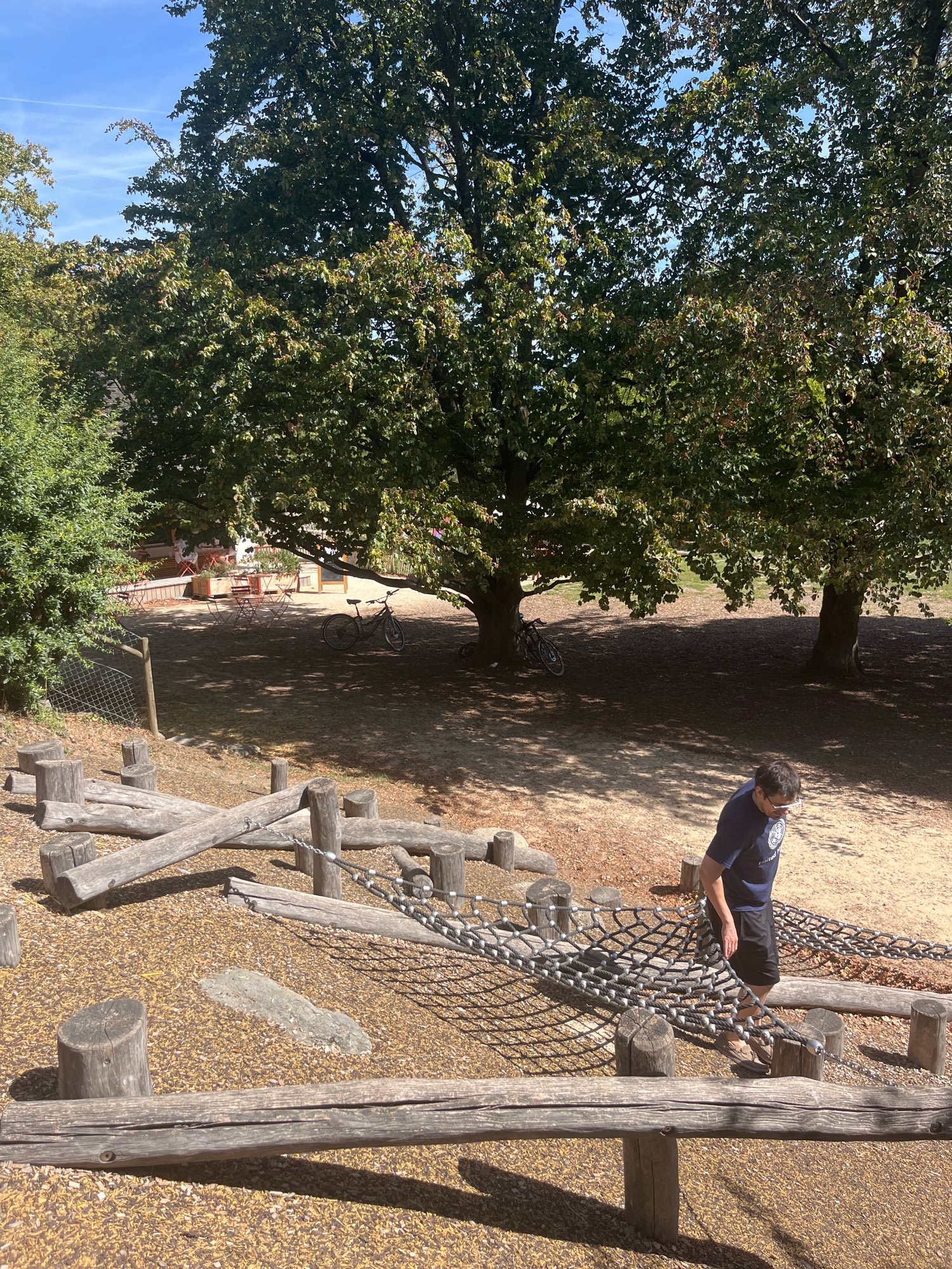 The image depicts an outdoor playground area with a focus on natural wood structures and vegetation. In the foreground, a series of wooden logs and beams form an obstacle course, with a rope net bridge spanning between some of the elements. A person, identified as a young male wearing a dark blue t-shirt and black shorts, is walking across the rope bridge. The background reveals a sandy area with trees providing shade, and a glimpse of a cafe or seating area with tables and chairs. Two bicycles are visible leaning against trees in the mid-ground. The sky above is a clear blue with a few wispy clouds. The scene is set on a sunny day. This playground appears to be located in Geneva, Switzerland.