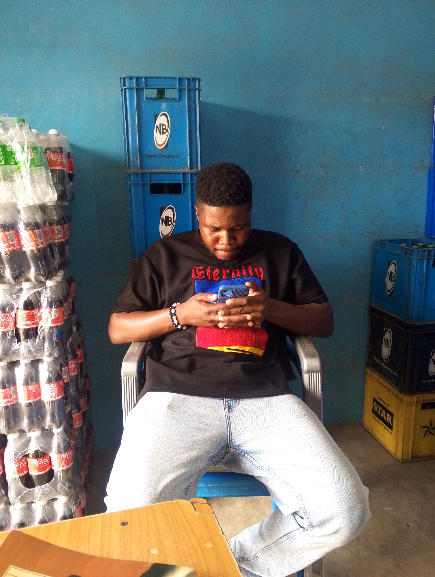 A man sits in a plastic chair, focused on his blue cell phone. He wears a black t-shirt with the word "Eternity" in red letters, blue jeans, and a bracelet. A stack of Coca-Cola bottles is to his left, while stacks of blue and yellow crates, labeled "NB" and "STAR", are on his right. A wooden desk is in the foreground. The setting appears to be a store or warehouse with a turquoise wall.