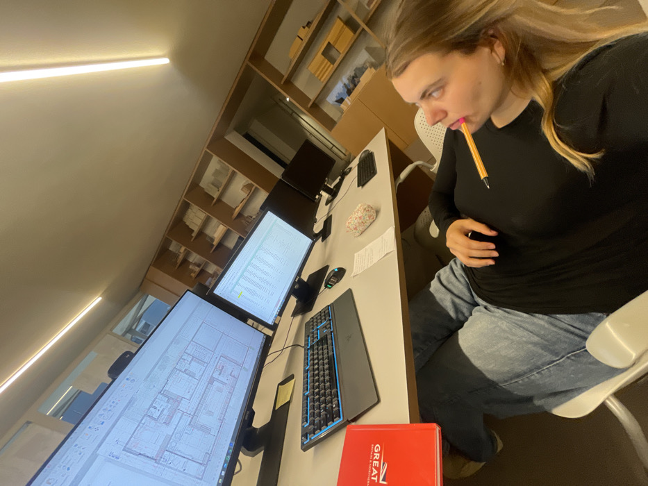 A female individual with light brown hair is seated at a white desk in a modern office environment in Mexico City, Mexico. She is observed from a slightly elevated, tilted perspective, looking upwards and to her right, with a yellow pen held horizontally between her lips. She wears a long-sleeved black top and blue jeans, with her left hand resting on her lap and her right leg crossed over her left.
The desk features a multi-monitor setup: a large left monitor displays an architectural floor plan, a central monitor shows a spreadsheet with rows of data, and a smaller, dark-screened monitor is positioned to the right. A dark mechanical keyboard with blue accents and a black computer mouse are positioned in front of the monitors. To the left, a red notebook or binder with "GREAT" visible on its cover rests on the desk. Other objects include a yellow sticky note on the base of the large monitor, a dark smartphone, a floral-patterned eyeglasses case, and a crumpled piece of paper.
The background consists of a long, light-wood shelving unit spanning the wall, filled with various items such as stacks of paper, small architectural models, and samples. Two linear LED light fixtures illuminate the ceiling. A reflective surface or window is visible in the upper left portion of the image.