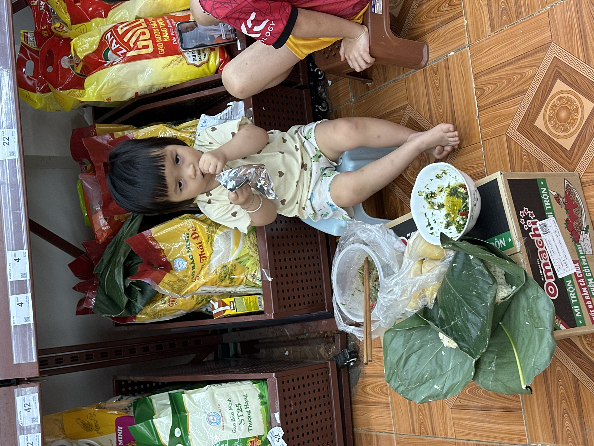 In Bắc Yên, Vietnam, a young Vietnamese child sits on a blue stool, eating from a foil packet. The child is wearing a light-colored t-shirt with heart patterns and light-colored shorts with cartoon characters. In front of the child, on a cardboard box, are two rice parcels wrapped in banana leaves, a plastic container of food with chopsticks, and a small bowl of sauce. The scene appears to be in a marketplace or a store, with shelves stocked with bags of rice in the background. To the right of the child, another person's legs are visible, suggesting the presence of another individual. The floor is tiled with a wooden pattern.