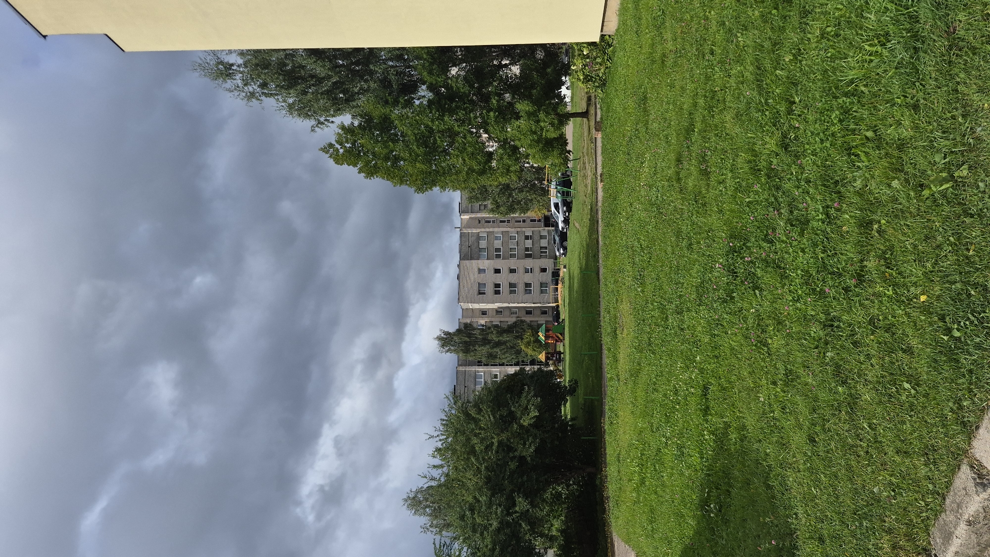 This is a full, eye-level shot of an apartment building in Krauja, Latvia, under a cloudy sky. In the foreground is a well-maintained, green lawn. Behind the lawn, a children's playground with colorful equipment is visible, with several cars parked nearby. The apartment building is a multi-story, concrete structure with many windows. Trees are scattered around the area, with a large, leafy tree on the right side of the frame and a bush in the middle-left. The overall impression is a typical urban residential area, with a focus on outdoor amenities and a somewhat overcast atmosphere.